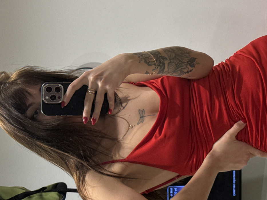 A woman with brown hair and bangs takes a selfie, holding her phone to obscure most of her face. She wears a red dress with thin straps. A dragonfly tattoo is visible on her chest. She has rings on her fingers and red nail polish. A floral tattoo is on her upper arm. A television is visible in the lower right corner of the frame.