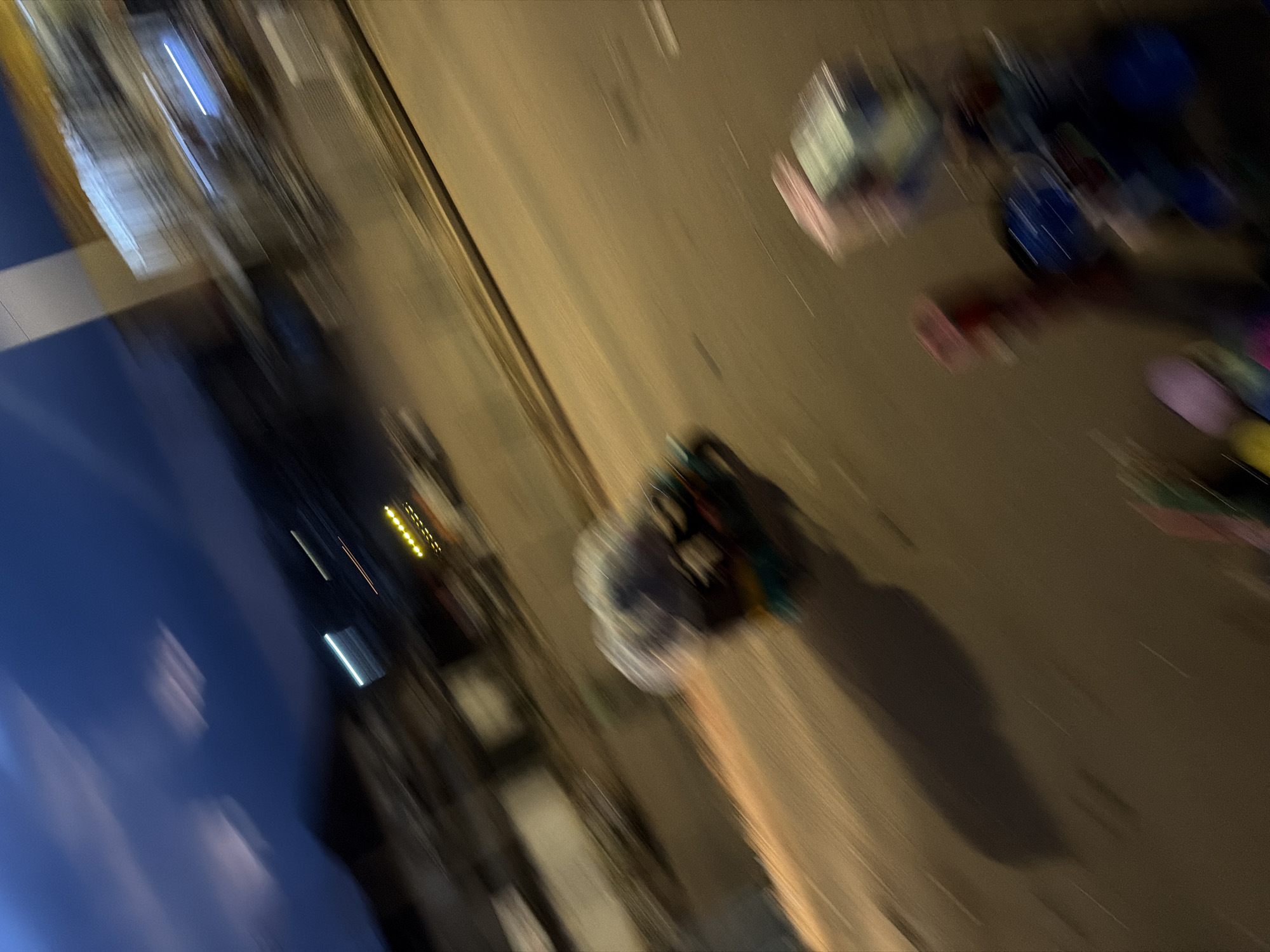 The image depicts a blurry, motion-blurred scene at night, suggesting movement. In the foreground, out-of-focus objects that appear to be brightly colored balloons or toys are visible. In the mid-ground, a blurry, multi-colored object that resembles a motorcycle or tricycle covered with a translucent tarp is moving. The background shows blurred buildings and lights, indicating an urban environment. The overall impression is of a street scene captured while in motion at dusk or night, possibly in Bukuru, Nigeria, given the context.