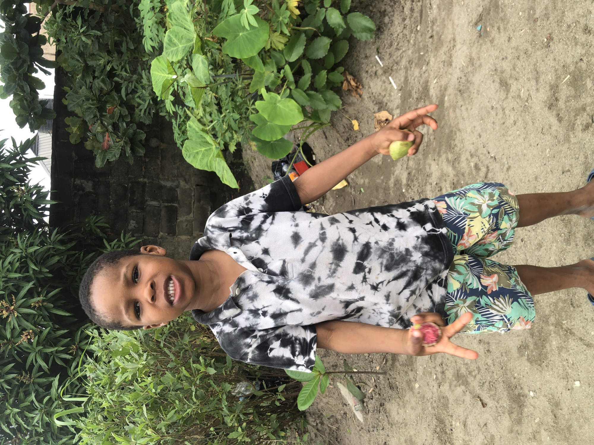 A young boy stands outdoors, facing the camera, with lush greenery and a brick wall in the background. The boy is wearing a tie-dye t-shirt with black and white patterns and floral-patterned shorts. He holds a small, light green fruit in his right hand and a reddish-pink fruit in his left hand, which is held in a V-shape between his index and middle fingers. The ground is sandy and uneven, with some scattered leaves and debris. The scene appears to be a residential area in Ado, Nigeria, characterized by the dense vegetation and visible construction elements.