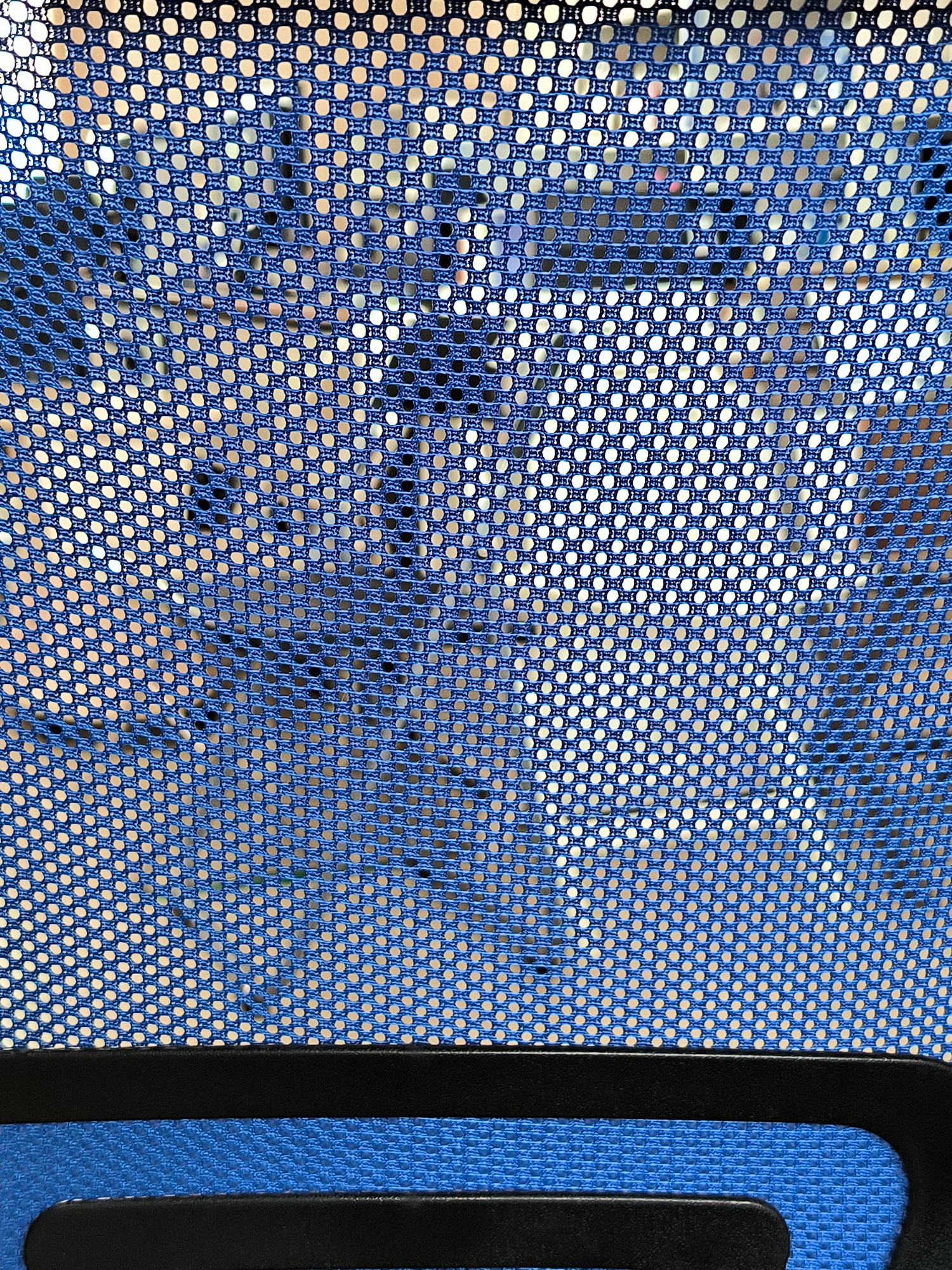 The image displays a close-up view of a blue mesh fabric, possibly from a chair or bag. The mesh has a regular pattern of small, circular holes. The fabric appears to be slightly worn or stretched in some areas, revealing a lighter background through the holes. At the bottom of the image, a black band and a blue textured surface are visible, suggesting this is part of a larger object. The lighting is diffused, creating a soft illumination of the fabric. There is no discernible information about the location in Yuzhno-Sakhalinsk, Russia, nor any people or specific actions.