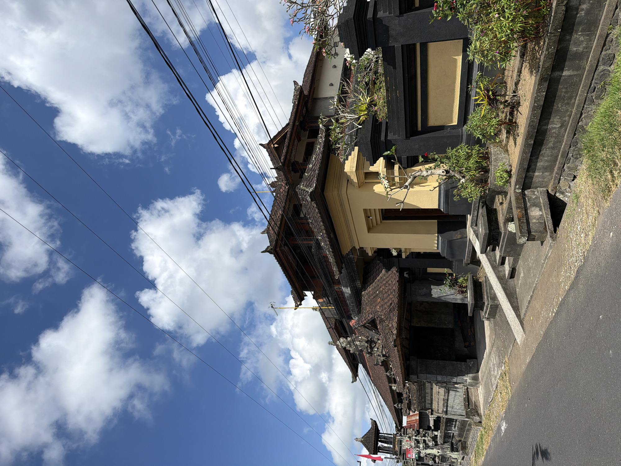 In the city of Petang, Indonesia, a traditional Balinese building stands under a bright blue sky dotted with fluffy white clouds. The building features a multi-tiered, dark tiled roof, ornate stone carvings, and a prominent yellow entranceway. A black and gold fence lines the front of the property, with a meticulously maintained garden featuring various plants and flowers. To the left, a small, ornate tower with a red flag is visible, suggesting the building might be a temple or a significant cultural site. Power lines crisscross the sky above the structure. The street in front is paved and appears to be a quiet road. No people are visible in the image.