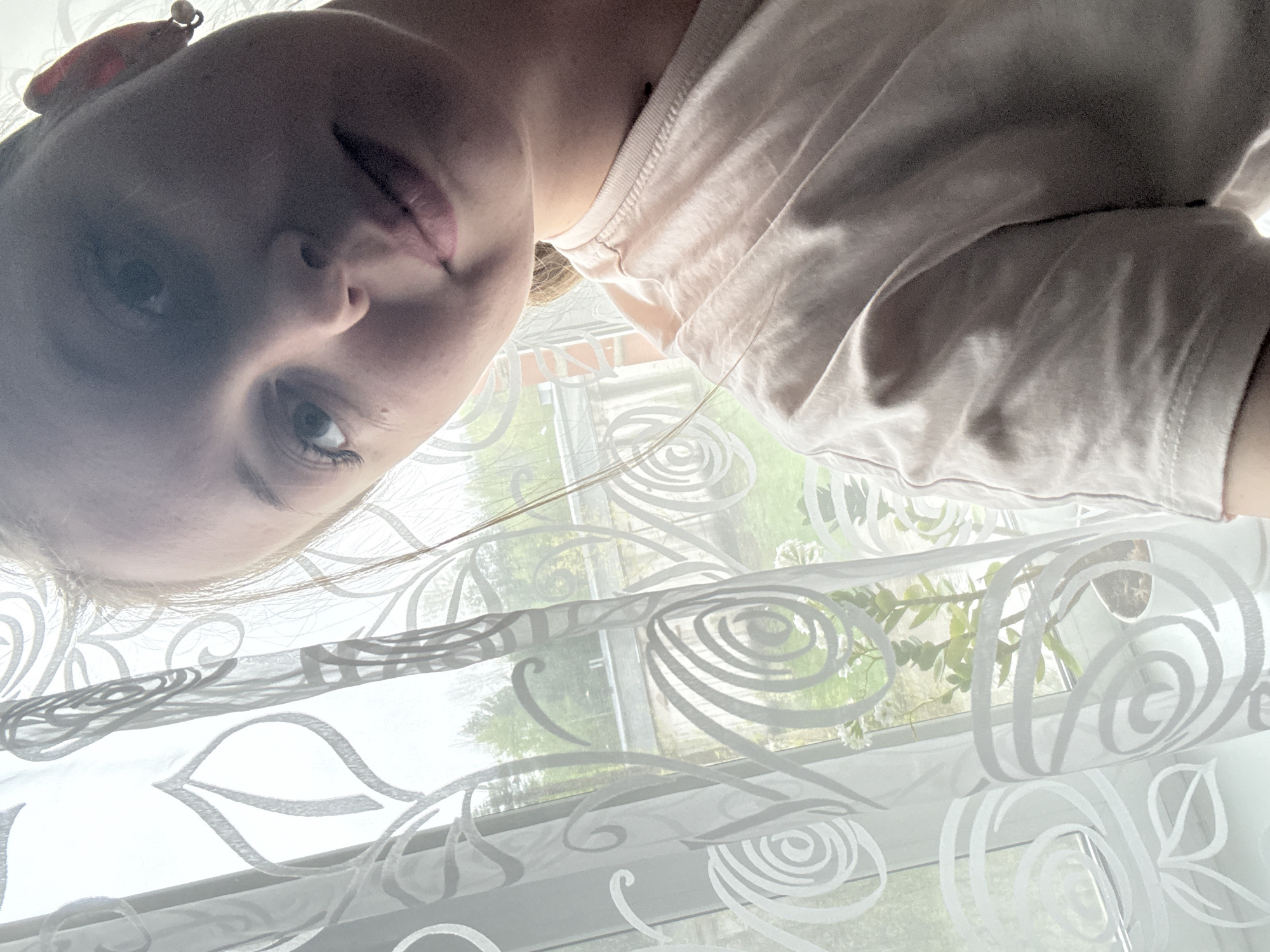 A close-up, slightly angled shot shows a person with fair skin and light-colored hair, positioned above what appears to be a window covered with a white, patterned curtain. The person's face is partially obscured by shadow, but their eye and part of their mouth are visible. They are wearing a light-colored shirt. Behind the curtain, a blurred view of a green, outdoor setting can be seen, possibly indicating trees or a garden. The location in the city of Unknown, Unknown, is not discernible.