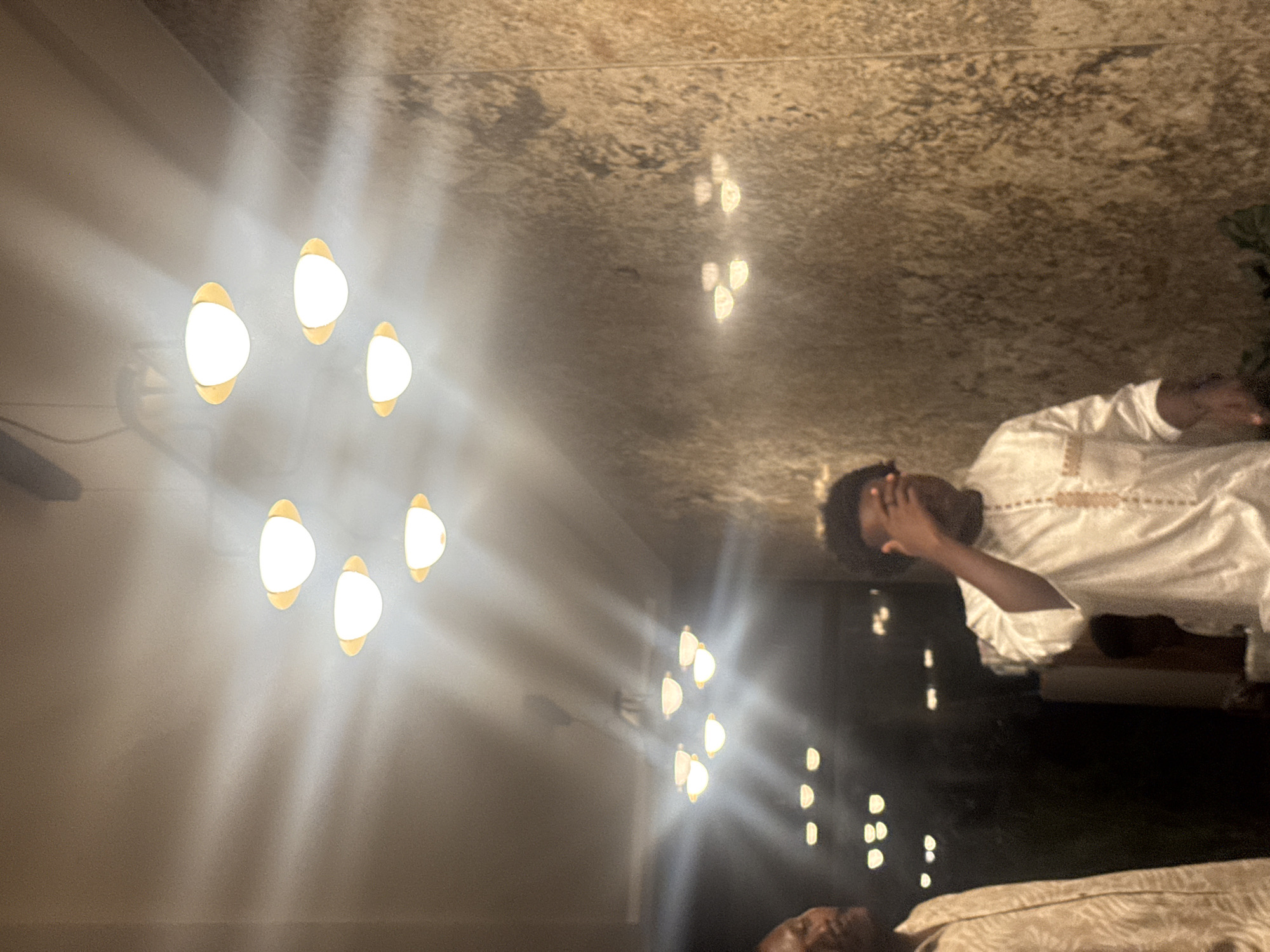 The media file shows an indoor scene with two individuals. One person is prominently featured, wearing a white traditional garment with embroidered details. They are partially obscured by their hand, which is covering their face, suggesting a possible emotion of distress, exhaustion, or reflection. The other person is in the foreground, also wearing a similar traditional garment, with their head and upper body visible.
The setting appears to be a dimly lit room, possibly a residence. Several modern-looking light fixtures are visible on the left side of the frame, emitting a warm, diffused glow that creates soft light patterns on the wall and ceiling. The ceiling has a textured, possibly stone or concrete, finish. The overall ambiance is subdued, with the lighting creating a somewhat dramatic and intimate atmosphere. The rotation of the image suggests it may have been captured in a vertical orientation and later rotated. There is no visible text or indication of the time of day or weather. The context of Abuja, Nigeria, suggests the traditional garments might be an Igbó or similar ethnic attire.