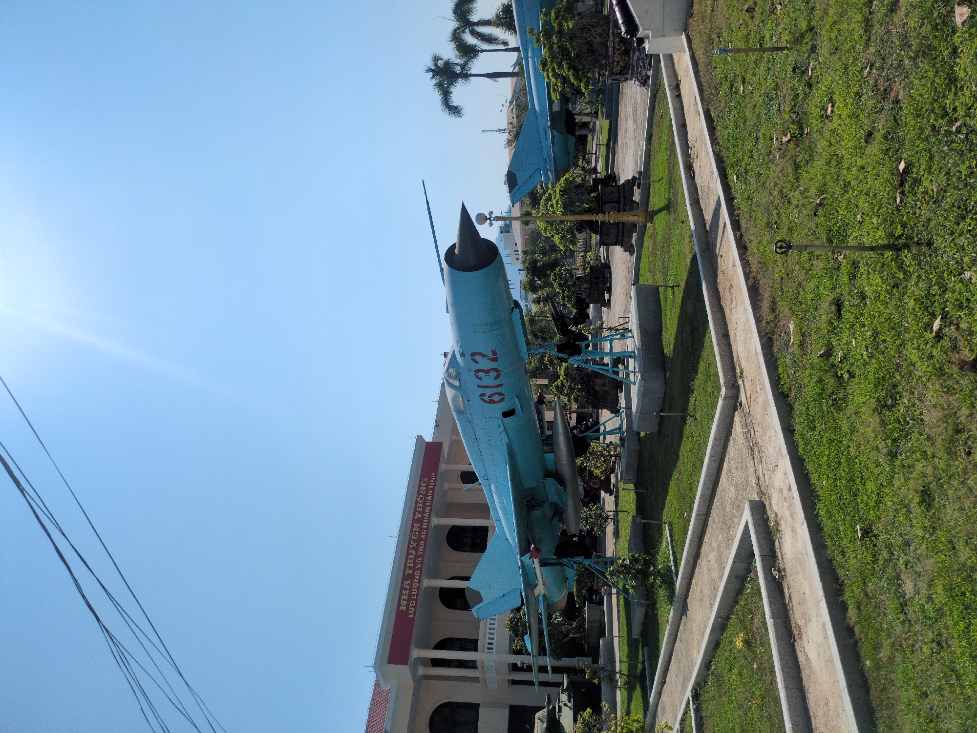A light blue MiG-21 fighter jet is displayed outdoors on a metal frame, with "6132" painted in red on its fuselage. The jet sits in a grassy area adjacent to a building with a red sign in Vietnamese. Additional military vehicles and artillery pieces are nearby. The background includes palm trees and a bright blue sky with power lines visible. The scene appears to be a historical military display or museum in South Quy Nhon Ward, Vietnam.