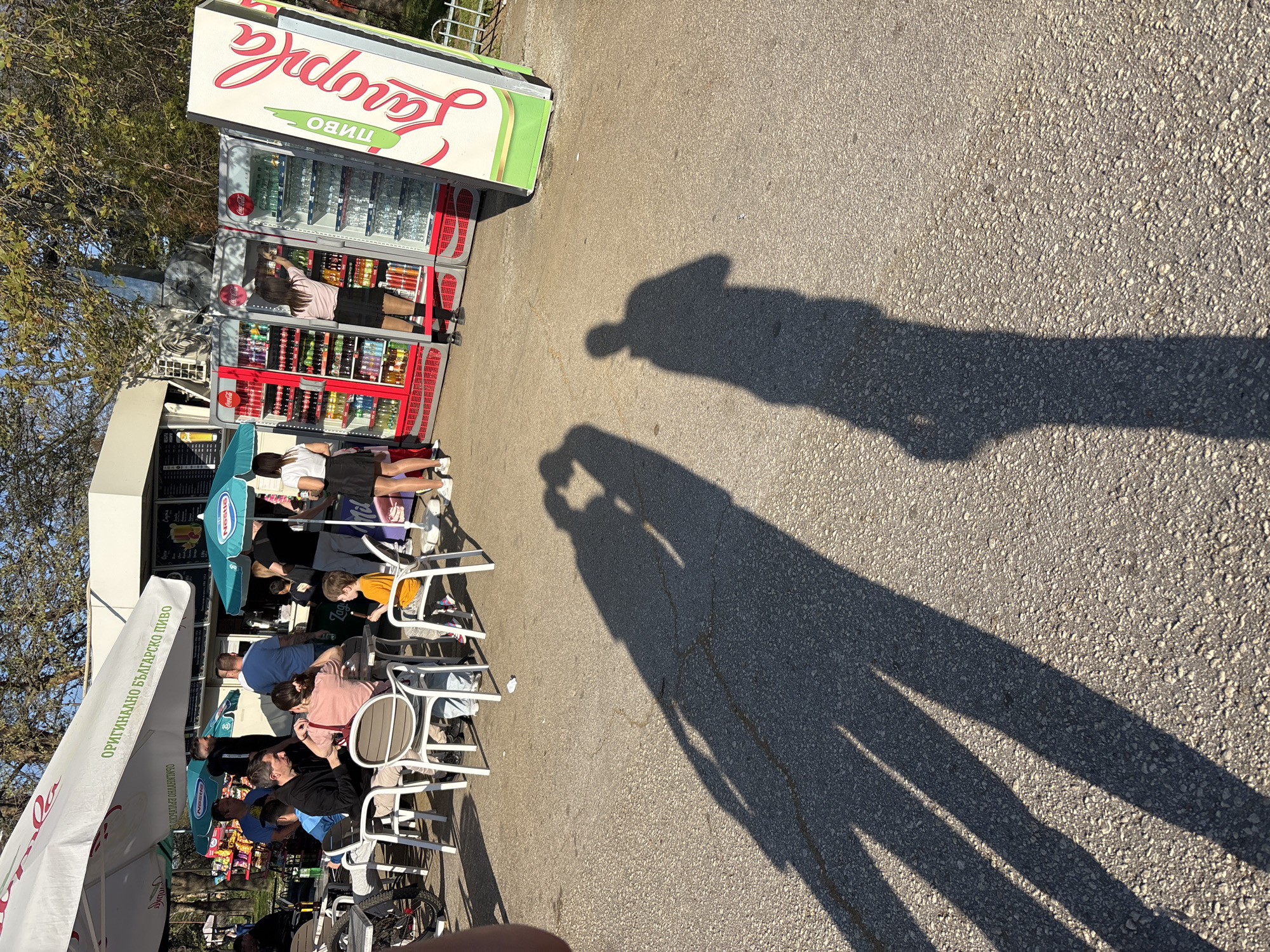 The image shows an outdoor cafe in Plovdiv, Bulgaria, on a sunny day. Several people are seated at white plastic tables and chairs under umbrellas, possibly eating or drinking. A refrigerated display case selling drinks like "Lavazza" and "пиво" (beer) is positioned near the cafe. Two tall shadows of people (likely the photographer and another person) stretch across the paved ground towards the cafe.