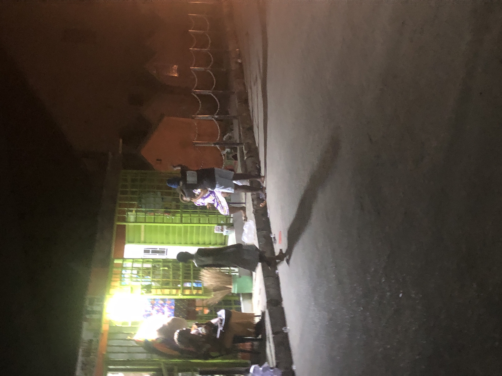 The image depicts a street scene in Ado, Nigeria, at night. The scene is lit by streetlights and lights emanating from businesses. Several people are visible along the street. One person is standing near the curb with a broom. A metal fence is behind the individuals. The street surface is asphalt. A ladder-like structure is set up against a building.