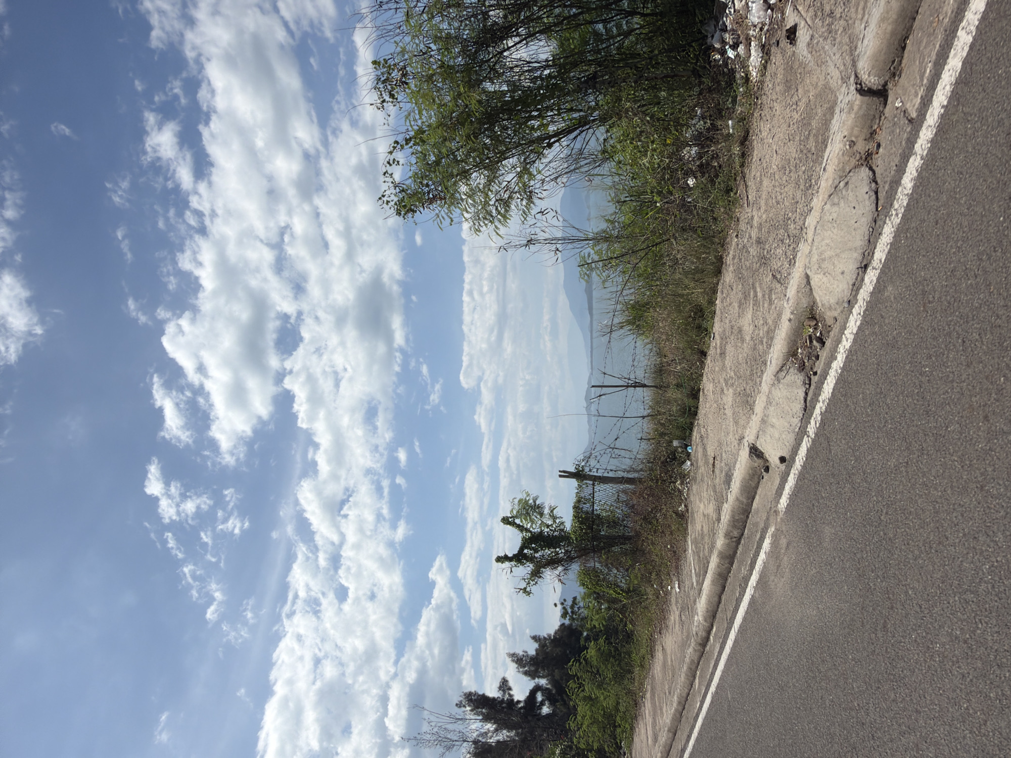 This media file shows a roadside view with a body of water and mountains in the background. The scene is dominated by a clear blue sky filled with fluffy white clouds. The foreground features a paved road with a white line marking its edge, and a concrete curb that appears worn and cracked in places. To the left of the road, there is a grassy embankment overgrown with various types of vegetation, including trees and bushes. Some litter is visible on the embankment. The background reveals a large body of water, possibly a bay or ocean, with mountains in the far distance. The overall impression is of a bright, sunny day, likely in the morning or afternoon, given the clear sky and good visibility. There is no activity or people depicted, suggesting a static shot of the landscape. The location is identified as Bac Nha Trang Ward, Vietnam, which aligns with the tropical vegetation and mountainous coastal scenery. No text is visible in the image.