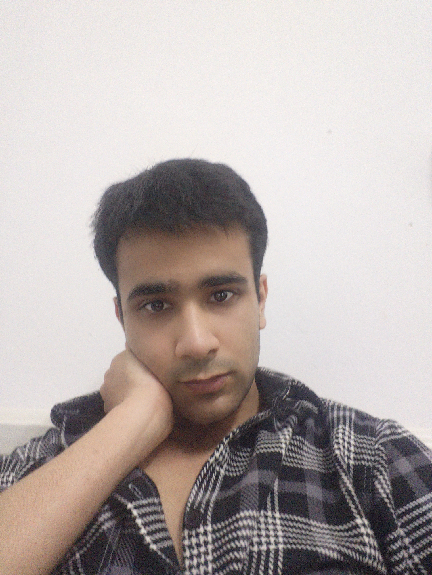 A young man is indoors, resting his chin on his hand. He has dark hair, dark eyes, and is wearing a black and white plaid shirt. The background is a plain white wall. The image does not provide specific details to confirm the location in Chongqing High-tech Zone, China.