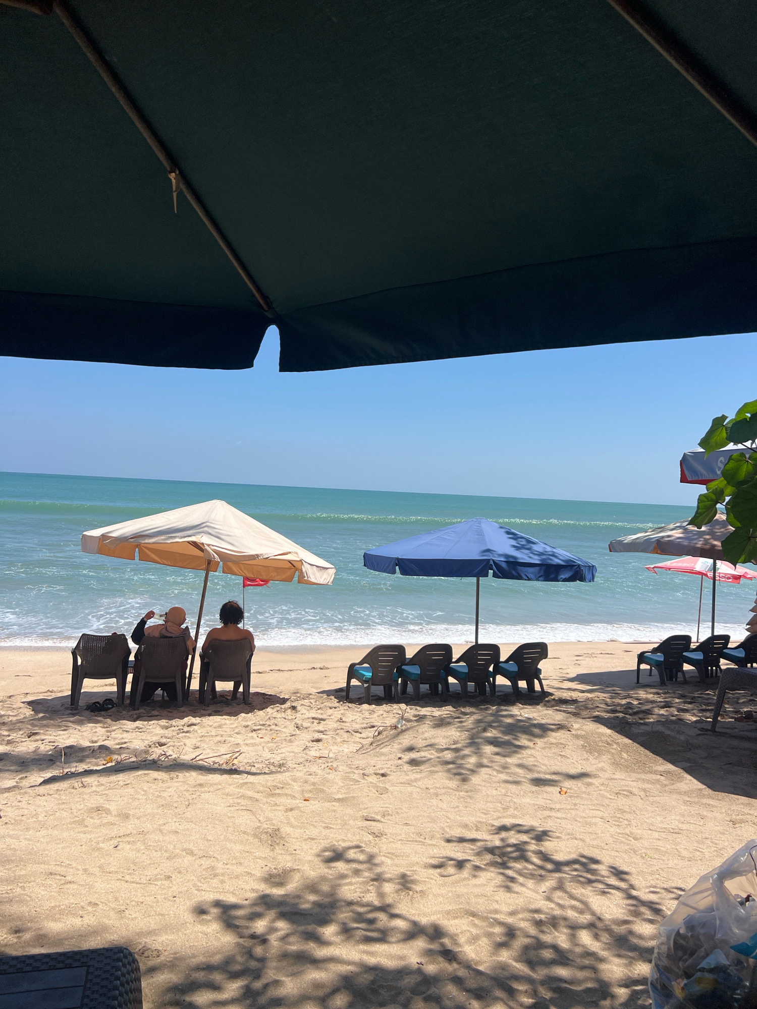 The image depicts a beach scene in Kuta, Indonesia, with a focus on a row of beach chairs and umbrellas set up on the sand. In the foreground, a dark green umbrella partially obscures the view. The middle ground shows the sandy beach with several beach chairs arranged in pairs, some occupied by people. Two individuals are seated under a beige umbrella, facing the ocean, with one appearing to be holding something. Another blue umbrella is positioned to their right, also with chairs beneath it. Further to the right, more beach chairs are visible, some with blue cushions. The background features the turquoise ocean with gentle waves, meeting a clear blue sky. The overall atmosphere is relaxed and summery.