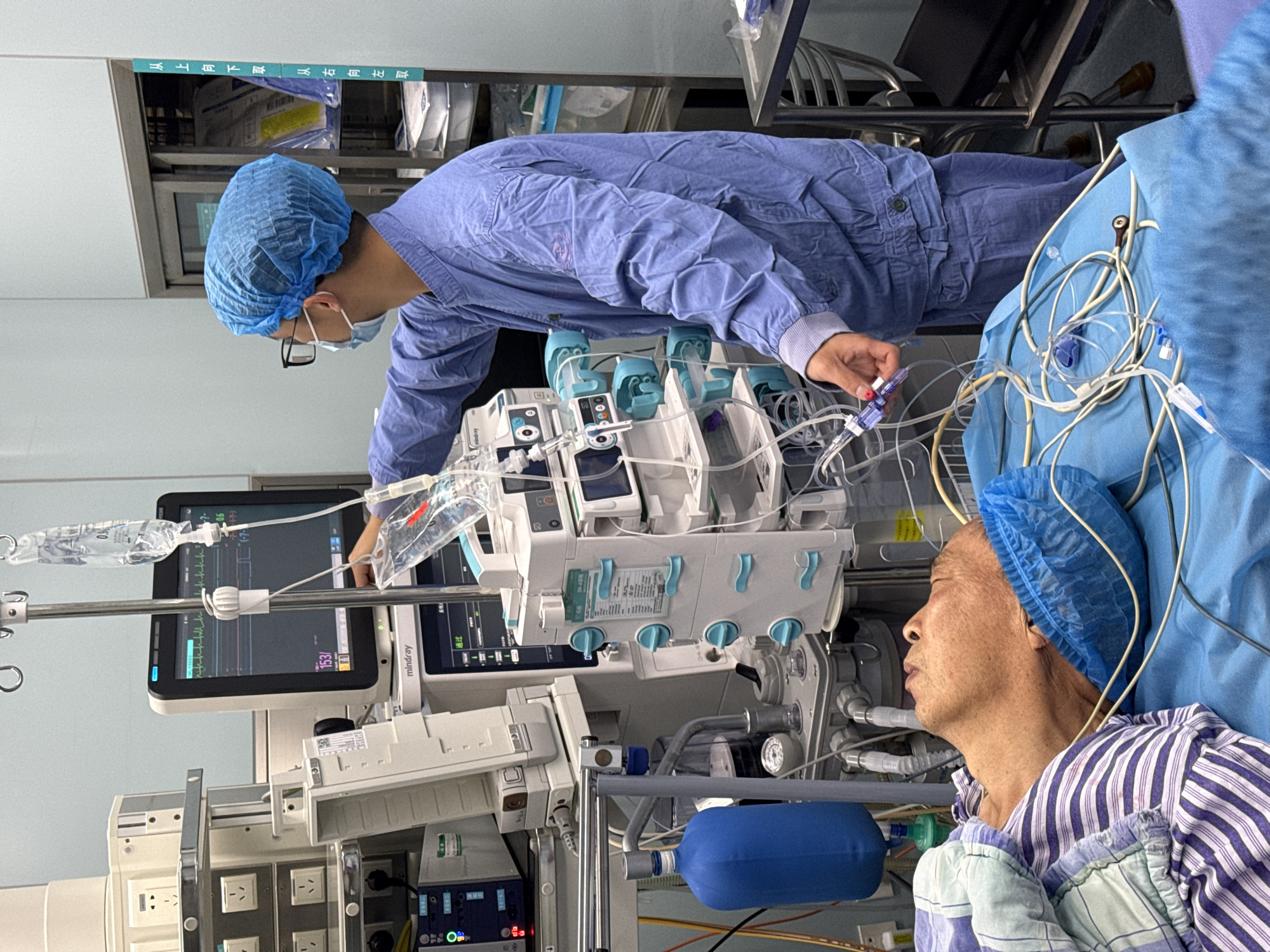 The image shows a patient in what appears to be a hospital setting. The patient is lying in a hospital bed, covered with a blanket with blue and white stripes, and wearing a blue surgical cap. Medical equipment surrounds the bed, including monitors, IV drips, and various tubes and wires connected to the patient. A medical professional, wearing a blue surgical cap and gown, is attending to the equipment. The scene suggests a medical procedure or monitoring in progress. Without additional information, the location in the city of Yuzhong District, China, cannot be independently confirmed.