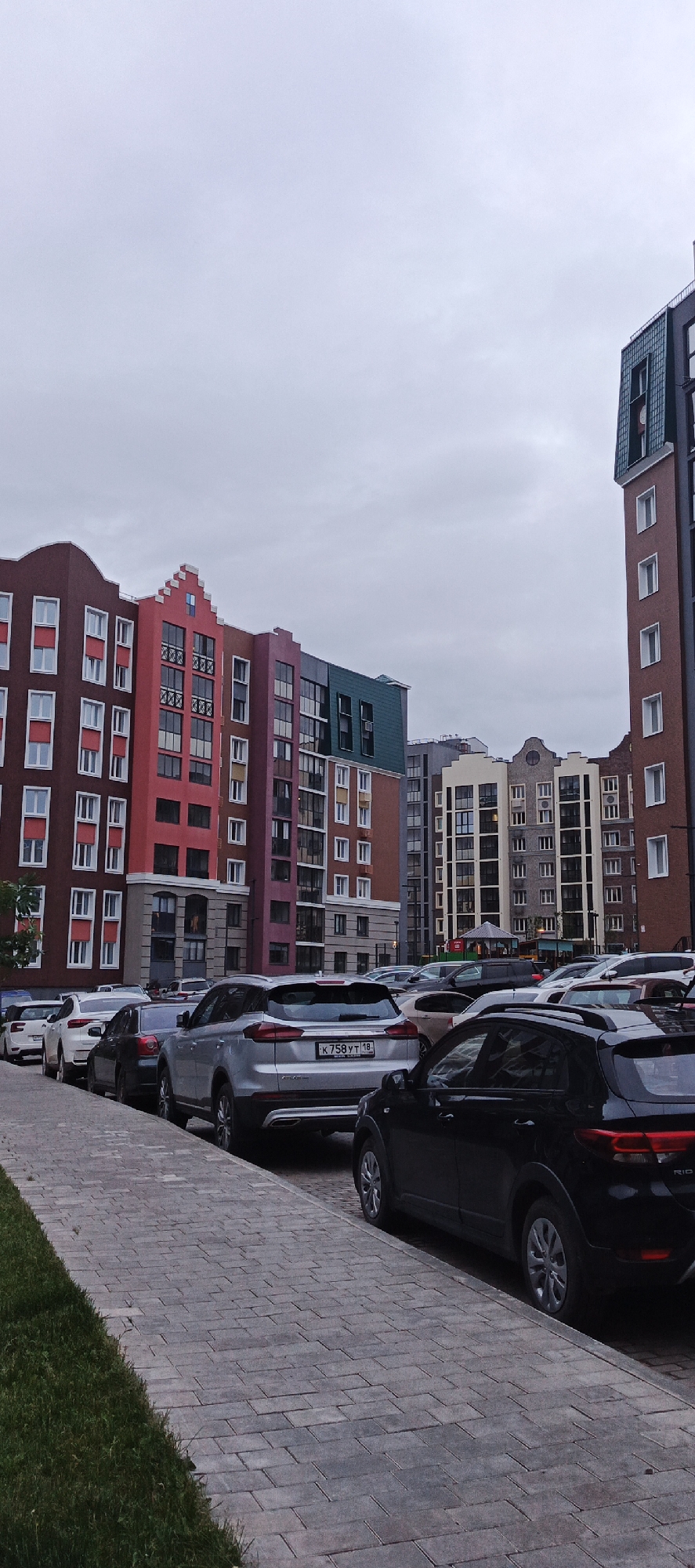 This image depicts an urban residential street scene under an overcast sky. The foreground features a grey and brown paver sidewalk with a narrow strip of green grass along its left edge. A line of vehicles is parked facing away from the camera along the right side of the sidewalk.
Prominent in the midground and background are multi-story apartment buildings exhibiting varied, colorful facades and distinct architectural styles, including gable roofs, stepped gables, and decorative pediments, reminiscent of Northern European or Dutch influence. From left to right, visible buildings include:
- A dark reddish-brown building with numerous white-framed windows.
- A red building featuring a prominent stepped gable with a small blue decorative element at its apex, along with balconies.
- A purple/mauve building transitioning to a dark teal roof or upper facade section.
- Further right, a building with a light beige or cream facade, grey architectural accents, and a rounded pediment with a central circular feature.
- On the far right, a tall, narrow, brown building with a steeply pitched dark teal roof section.
Multiple cars are parked on the street. The most clearly identifiable vehicles from left to right are:
- A white sedan.
- A dark grey sedan.
- A silver SUV, likely a Geely Atlas, displaying a Russian license plate "K 758 YT 18". The "18" indicates the Udmurt Republic region.
- A black compact SUV, with a partial "RIO" badge visible on its rear.
Additional cars are visible further down the street and in a parking area behind the initial row.
No individuals are visible in the scene. The sky is a uniform light grey, indicating full cloud cover. The overall environment appears to be a modern residential development within an urban setting. Based on the provided information, the location is within Pervomaysky, Russia.