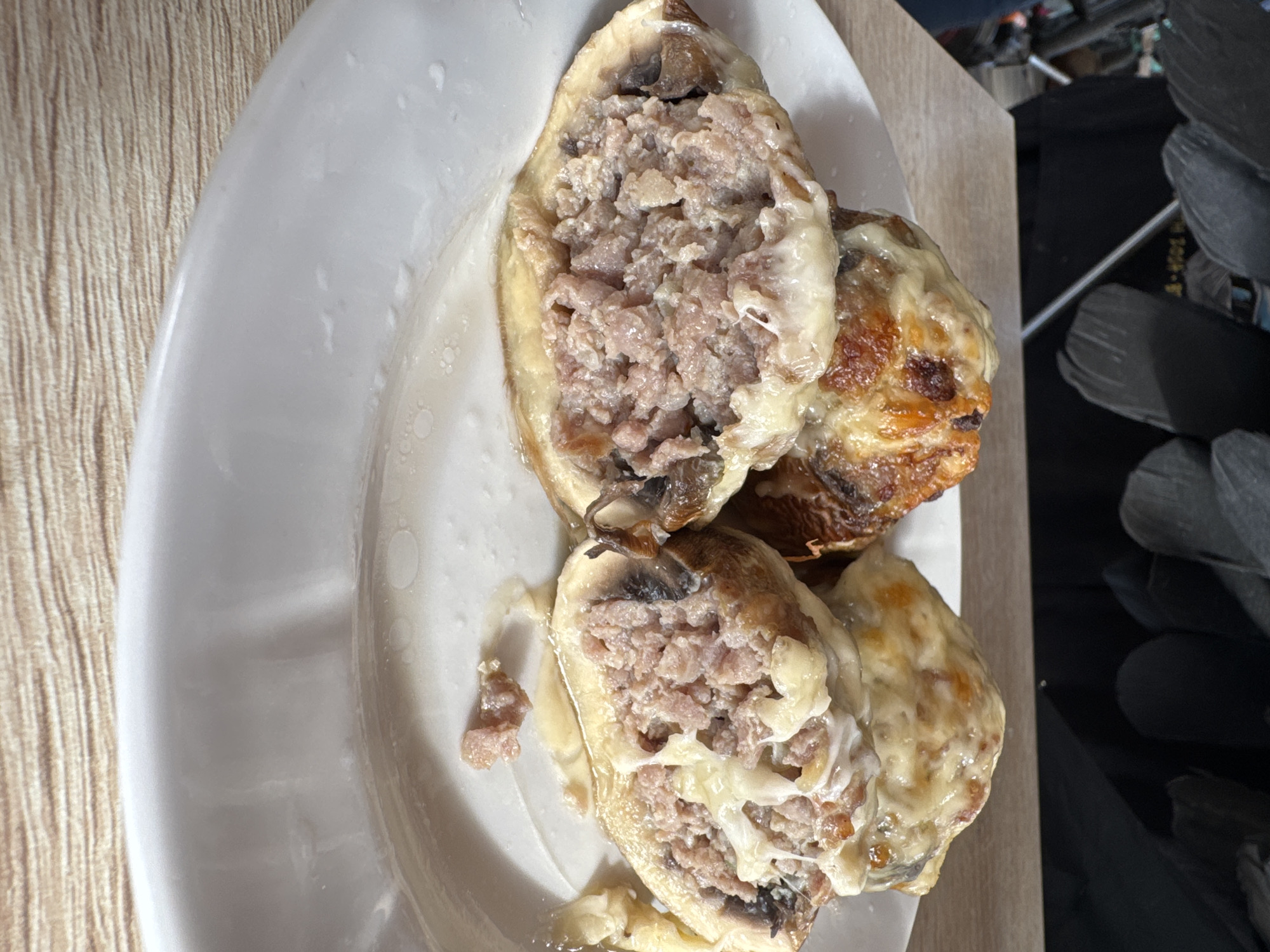 The image depicts a white plate with three stuffed mushrooms. The mushrooms are filled with a ground meat mixture, possibly pork or beef, and topped with melted cheese. The mushrooms appear to be baked, with some browning on the cheese. The plate is placed on a wooden surface, possibly a table or countertop. There is an object on the right side that appears to be either chair or cleaning tool. The location and city of Chaikovsky, Russia cannot be confirmed.