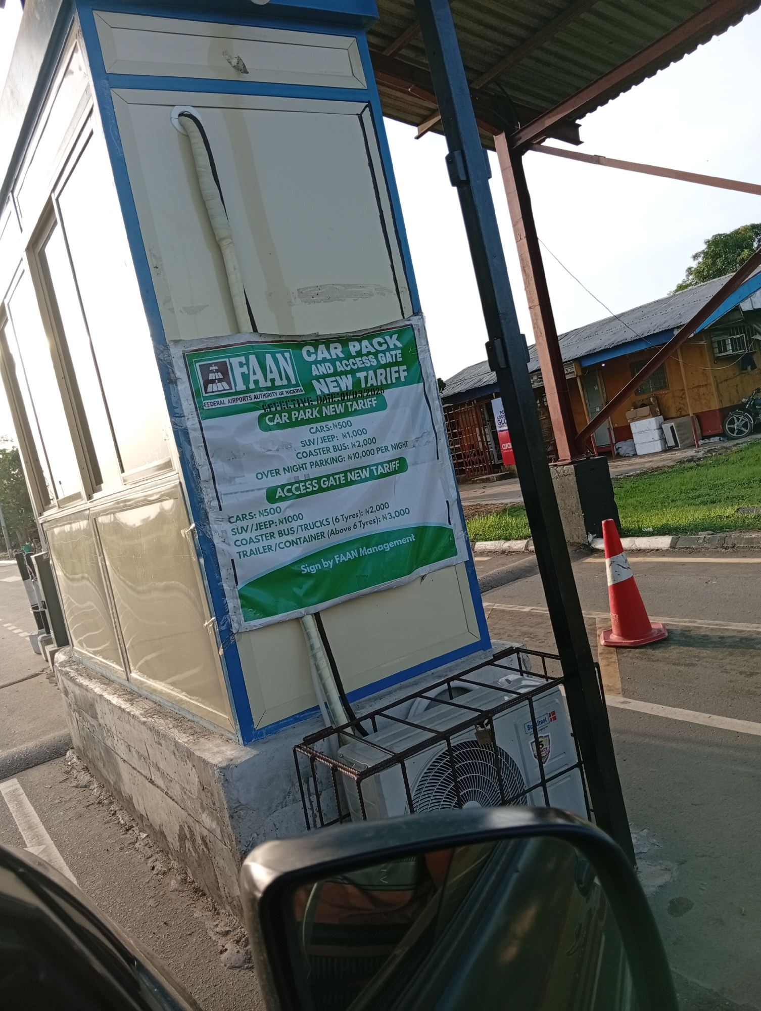 The image shows a parking and access gate booth, seemingly at an airport facility in Omagwa, Nigeria. A sign is affixed to the booth that details the "CAR PACK AND ACCESS GATE NEW TARIFF" for various vehicles. The sign indicates "FAAN" (Federal Airports Authority of Nigeria) as the managing entity. The booth itself is painted in off-white with blue trim. An air conditioning unit is placed outside the booth in a protective cage. Behind the booth is a partial view of a building, some greenery, and a safety cone. A car side mirror is in the foreground.