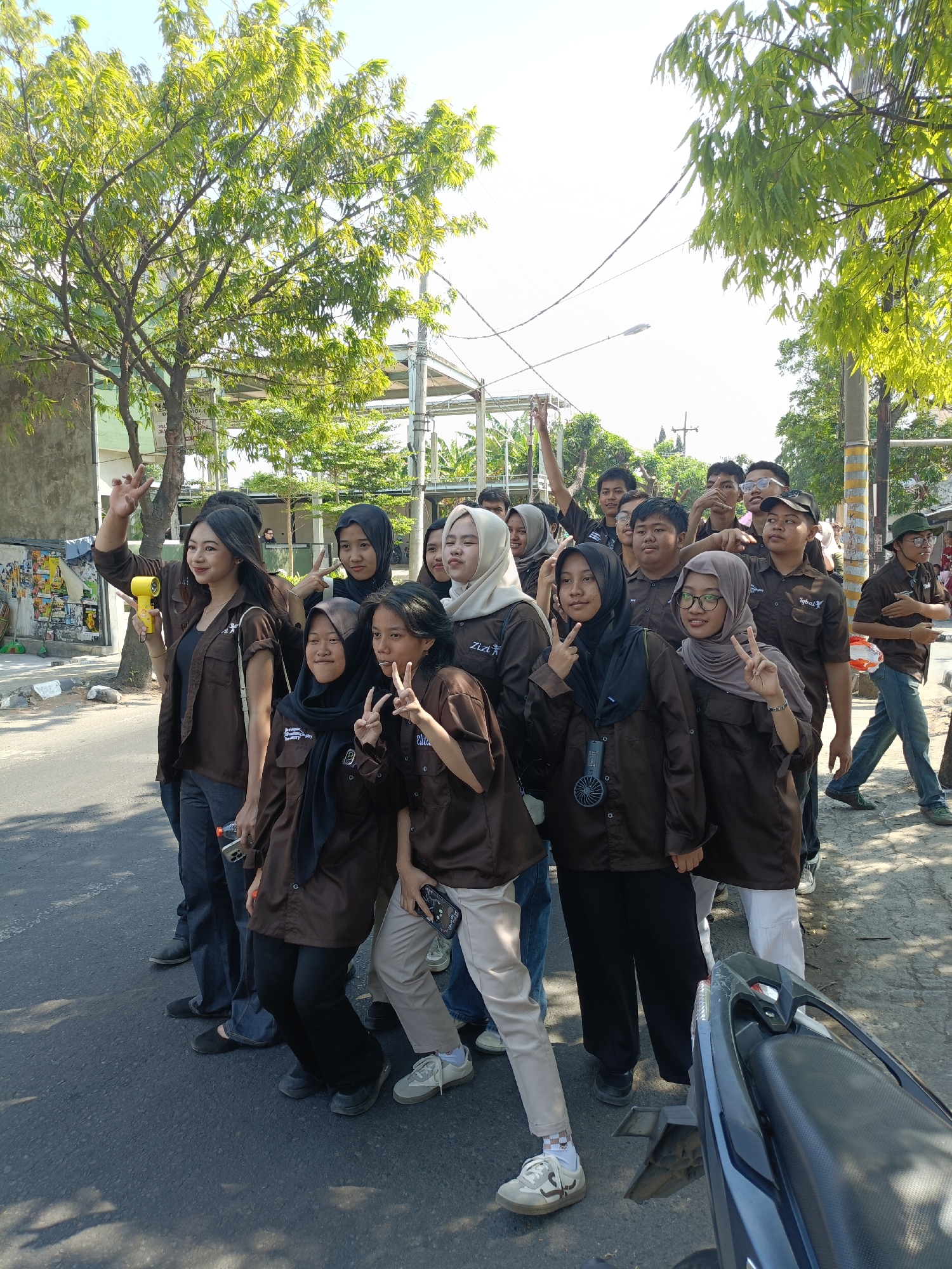 A group of approximately 15 young individuals, predominantly wearing matching brown shirts and some with headscarves, are posing for a photo outdoors. The scene appears to be on a street in a city, with trees lining the right side and buildings visible in the background. The lighting suggests it is daytime. The individuals are interacting and smiling, with several making peace signs with their fingers. One person in the foreground is holding a yellow object that resembles a small megaphone or spray can. A motorcycle is partially visible in the foreground on the right. The environment includes a paved road, a sidewalk, and a concrete wall on the left with some posters. The overall atmosphere is casual and social.