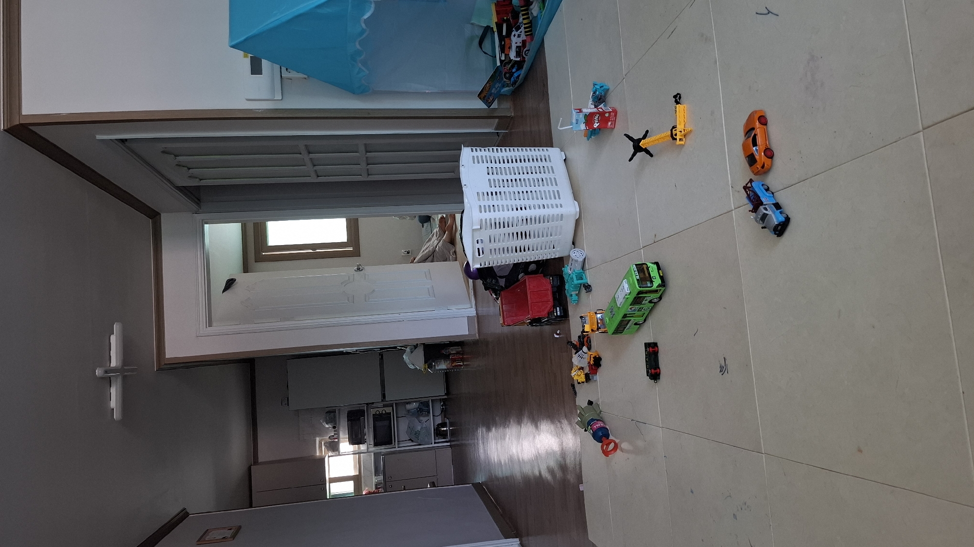 The image depicts a cluttered indoor scene, likely a child's play area within a residential dwelling in Daeso, South Korea. The floor is tiled and strewn with various toys, including several toy vehicles: a green bus, a red dump truck, a tow truck, and an orange sports car. A white plastic laundry basket filled with items is centrally located, and a blue play tent is visible to the right. In the background, doorways lead to other rooms, revealing glimpses of furniture, appliances, and a person lying on the floor. The lighting suggests it is daytime.