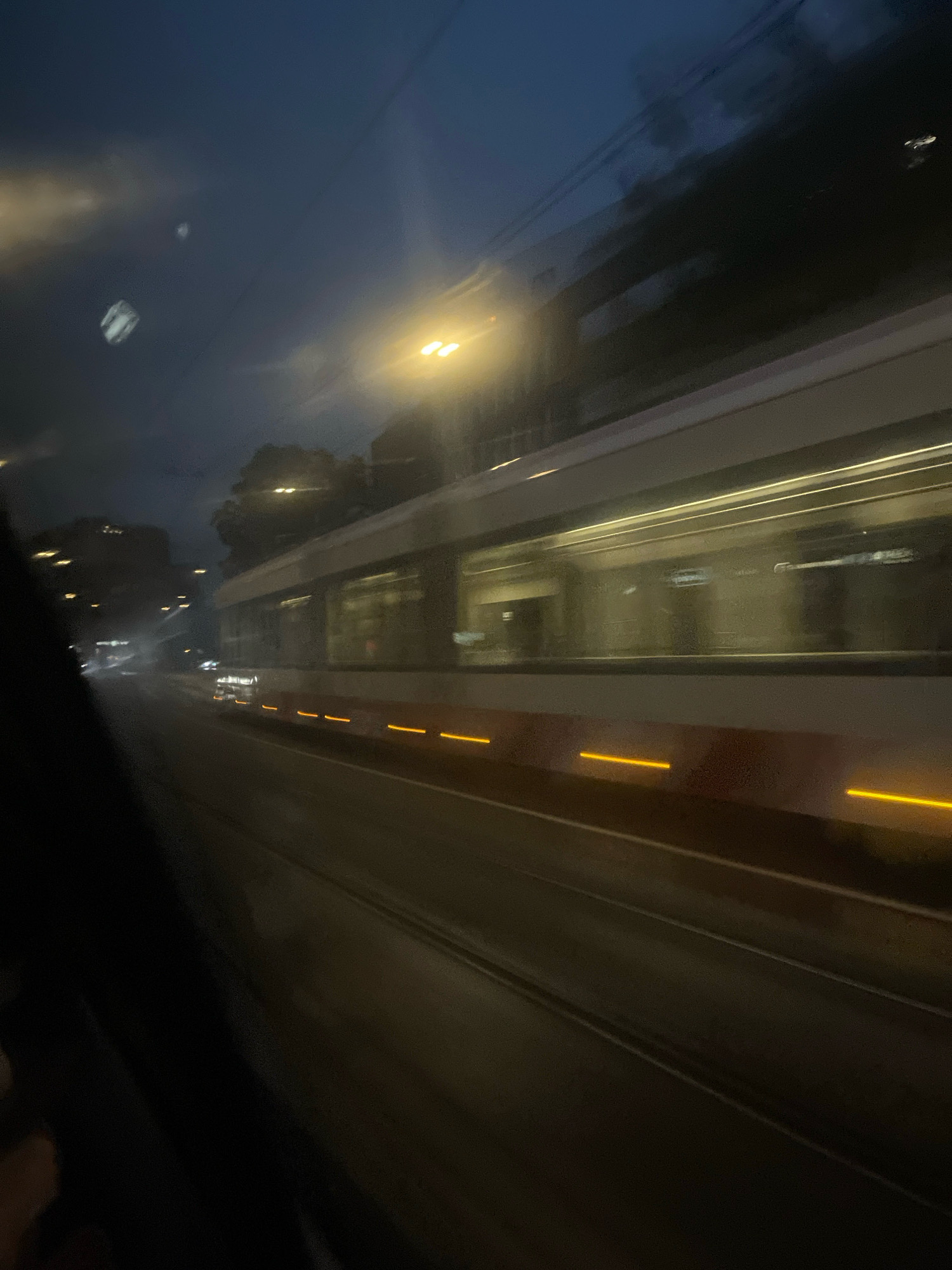 The image depicts a blurry view from inside a vehicle, likely a tram, as it travels through Tallinn, Estonia, during the evening. The sky is dark blue, with some faint lights visible. The tram itself is a prominent subject, with its white and red exterior and illuminated windows suggesting interior lighting. Yellowish lights are seen along the side of the tram and on the tracks. The motion blur indicates movement, and the overall atmosphere is dim and nocturnal. Buildings and trees are visible in the distance, also blurred by the movement. The scene captures a typical urban nightscape in Tallinn, characterized by the illuminated tram and the surrounding twilight environment.