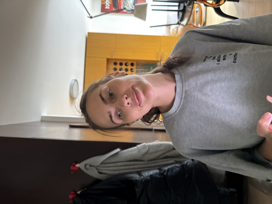 This image depicts a female subject in an indoor residential setting. The specific location within Warsaw, Poland, cannot be determined from the visual information provided.
The subject, a woman with light to medium brown hair gathered at the back, possesses fair skin and light-colored eyes. Her head is tilted slightly to her right, and she displays a subtle, closed-mouth smile while looking upwards and to the viewer's right. She is wearing an oversized grey long-sleeved sweatshirt. Text is present on the left chest area of her sweatshirt; partially visible segments include the words "my life,", "I didn't", "want to", and "come". Her right hand is partially visible at the bottom of the frame, holding a white-cased smartphone, with an index finger positioned as if interacting with the screen.
The background on the left features a dark brown wall with two distinctive red, stylized wall hooks. A black puffy coat hangs on the left hook, and a light grey coat or robe hangs on the right hook.
To the right of the subject, a brighter room is visible, separated by a white wall segment or doorway. This room contains yellow wooden cabinetry; one section of which displays a pattern of circular cutouts, possibly for bottle storage. A round, flush-mount ceiling light fixture is visible on the white ceiling. Further to the right, a black adjustable floor lamp stands next to a wall featuring a red and black framed poster or artwork. Two chairs, one black with thin legs and another with an orange-brown seat, are inverted and stacked on a table in the background. The flooring appears to be light-colored wood. The environment is illuminated by ambient light, suggesting an interior space.