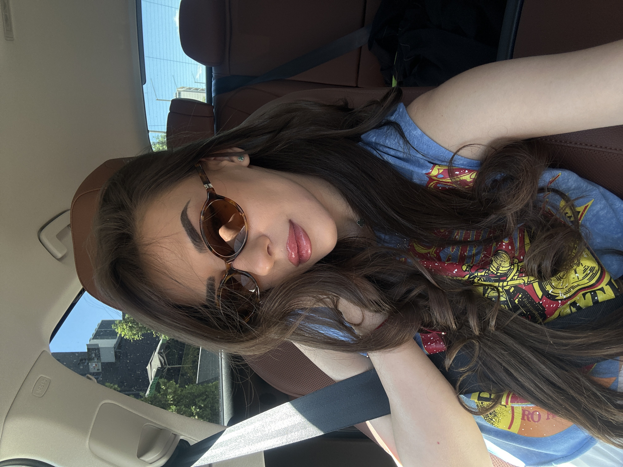 A young woman with long, dark, wavy hair is captured in a selfie taken from inside a car. She is wearing tortoiseshell sunglasses and a blue t-shirt with a colorful graphic print. Her lips are glossy, and she has light makeup. The interior of the car features brown leather seats and a seatbelt across her chest. Through the car window, an urban landscape is visible, including buildings and trees. The scene appears to be in Sofia, Bulgaria, based on the context provided.