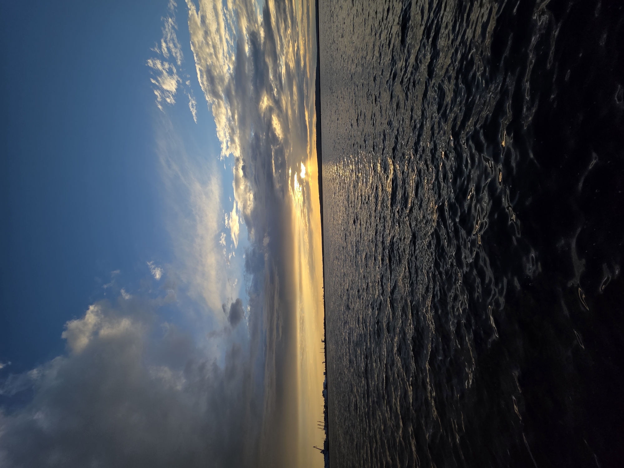 This media file depicts a scenic view of a body of water at sunset, with the horizon dividing the sky and the water. The sky is filled with a dramatic display of clouds, ranging from bright white to darker grays, illuminated by the warm, golden hues of the setting sun. The sun itself is partially obscured by clouds, casting a radiant glow across the sky. The water is dark and textured, with ripples catching the light, reflecting the colors of the sky and creating streaks of golden shimmer. In the distance, along the horizon, a faint silhouette of what appears to be a shoreline with industrial structures, possibly a port or city skyline, is visible. The overall mood conveyed is one of natural beauty and tranquility, characteristic of a sunset over a large body of water, likely a river or a bay. The time of day is clearly evening, as indicated by the sunset colors and lighting. The location is identified as Chaikovsky, Russia, which suggests a likely setting of a Russian river or reservoir. There is no visible text in the image. The composition is split vertically, with the sky occupying the left half and the water the right half.