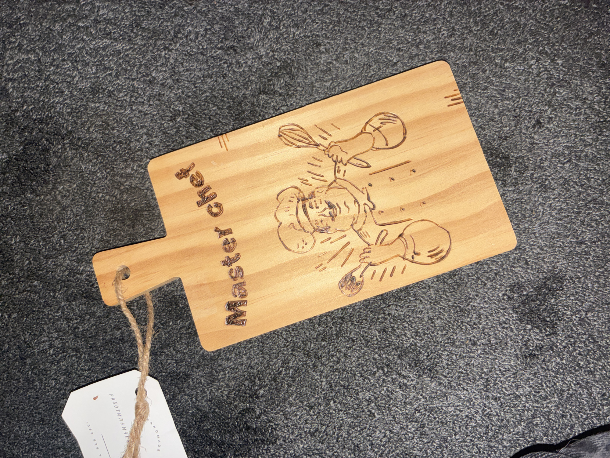 A wooden cutting board with "Master chef" etched into it lies on a dark grey, fuzzy surface, possibly carpet. The cutting board features an engraved illustration of a chef holding two kitchen utensils. The scene is indoors, with no visible indicators of a specific city or location. A white tag is attached to the cutting board by a piece of twine.