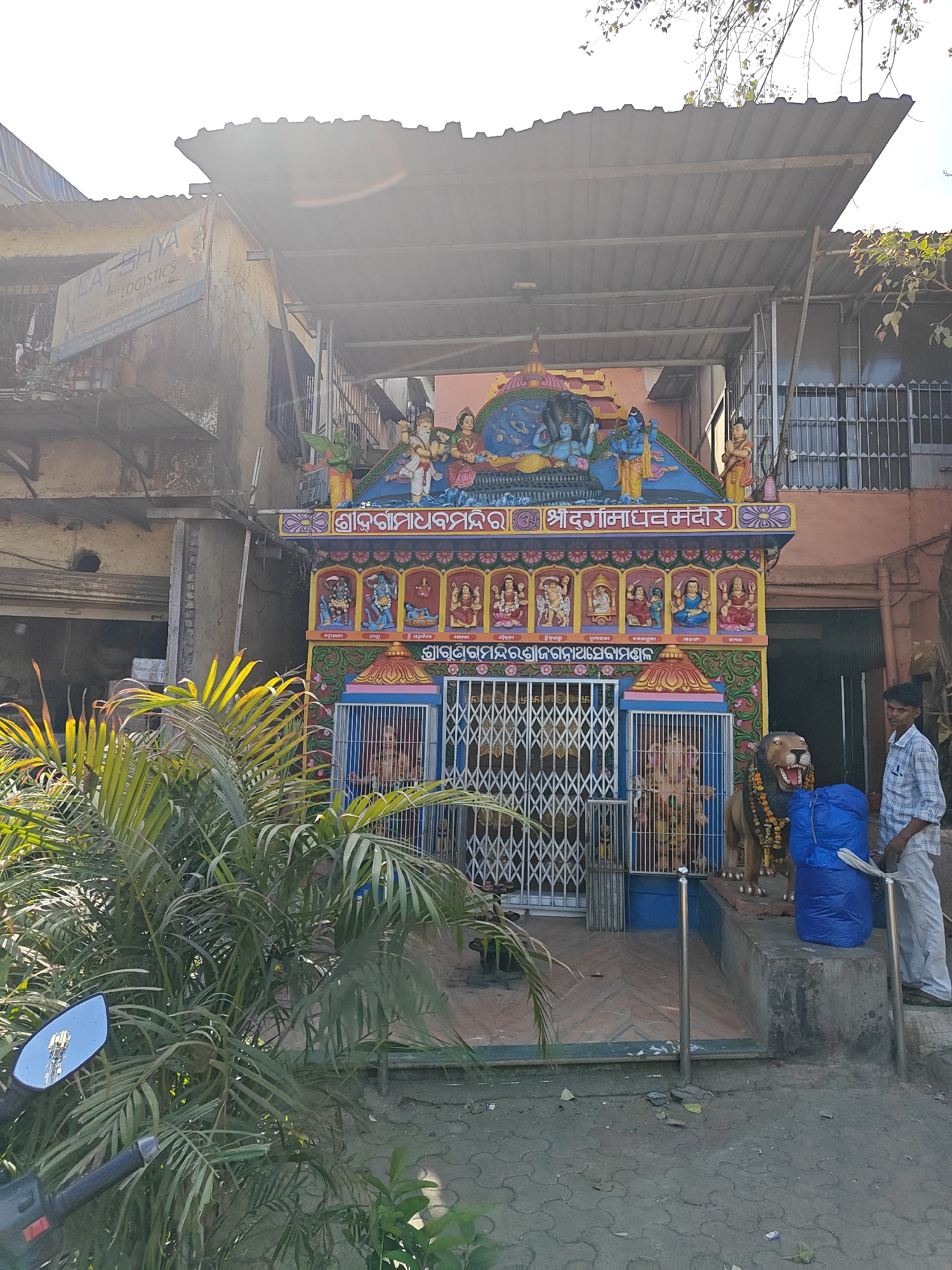 Here is a detailed description of the image:
**Overview:**
The image depicts a small, brightly colored Hindu temple situated in an urban environment, likely in Mumbai, India. A man stands next to the temple.
**Temple Details:**
The temple is adorned with multiple vibrant colors and intricate designs. It features representations of Hindu deities and religious symbols. Above the entrance are colorful figures and inscriptions in a local language (possibly Marathi or Hindi). The structure has a metal roof or awning providing shelter. A metal gate encloses the temple's interior. A statue of a lion decorated with garlands is placed next to the temple entrance.
**Surroundings:**
The temple is flanked by buildings, with one showing signs of age and wear. A sign board with the word 'Logistics' is visible on the upper left of the building behind the temple. The foliage of a potted plant partially obscures the view of the left side of the temple.
**People:**
A man stands to the right of the temple, near the base of the steps, wearing a light-colored shirt and pants. He is holding what appears to be some white rags. A blue tarpaulin is placed nearby on the temple steps.
**Overall Impression:**
The image captures a blend of religious devotion and urban life, with the temple serving as a focal point within a densely populated area. The man's presence suggests the temple is still actively used for religious practice. The presence of logistics company indicates an commercial environment.