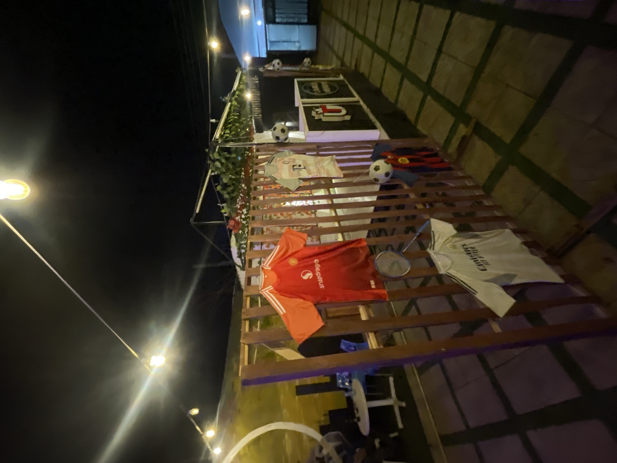 This image captures a sports-themed outdoor or semi-outdoor space at night in Bar Jirgi Summa, Nigeria, illuminated by numerous string lights. The scene is dominated by a wooden slatted structure, possibly a wall or overhead trellis, adorned with various items.
Several football (soccer) jerseys are prominently displayed on the wooden slats. One is a red and orange jersey with white sleeves, featuring the "snapdragon" logo and text. Another is a white long-sleeved jersey with the text "Emirates FLY BETTER" visible on the front. A light grey or white patterned jersey is also visible, along with a partially obscured dark red/black striped jersey. Multiple white and black footballs are placed among the jerseys and on the structure. A badminton racket rests next to the red and orange jersey.
To the left, green foliage and possibly artificial plants are draped along a metal framework. In the background, elevated on a platform, are two framed signs or displays; one appears to have the text "JUMANJI" (or similar lettering) and another features a stylized "U" logo. The ground below the structure has a patterned surface, resembling a grid of light-colored squares with dark lines, possibly tiles or painted concrete. The lighting casts a warm glow and creates reflections, emphasizing the nighttime setting. No people are visible in the image, suggesting the focus is on the decorated space itself, likely a bar or lounge area designed for sports enthusiasts.
