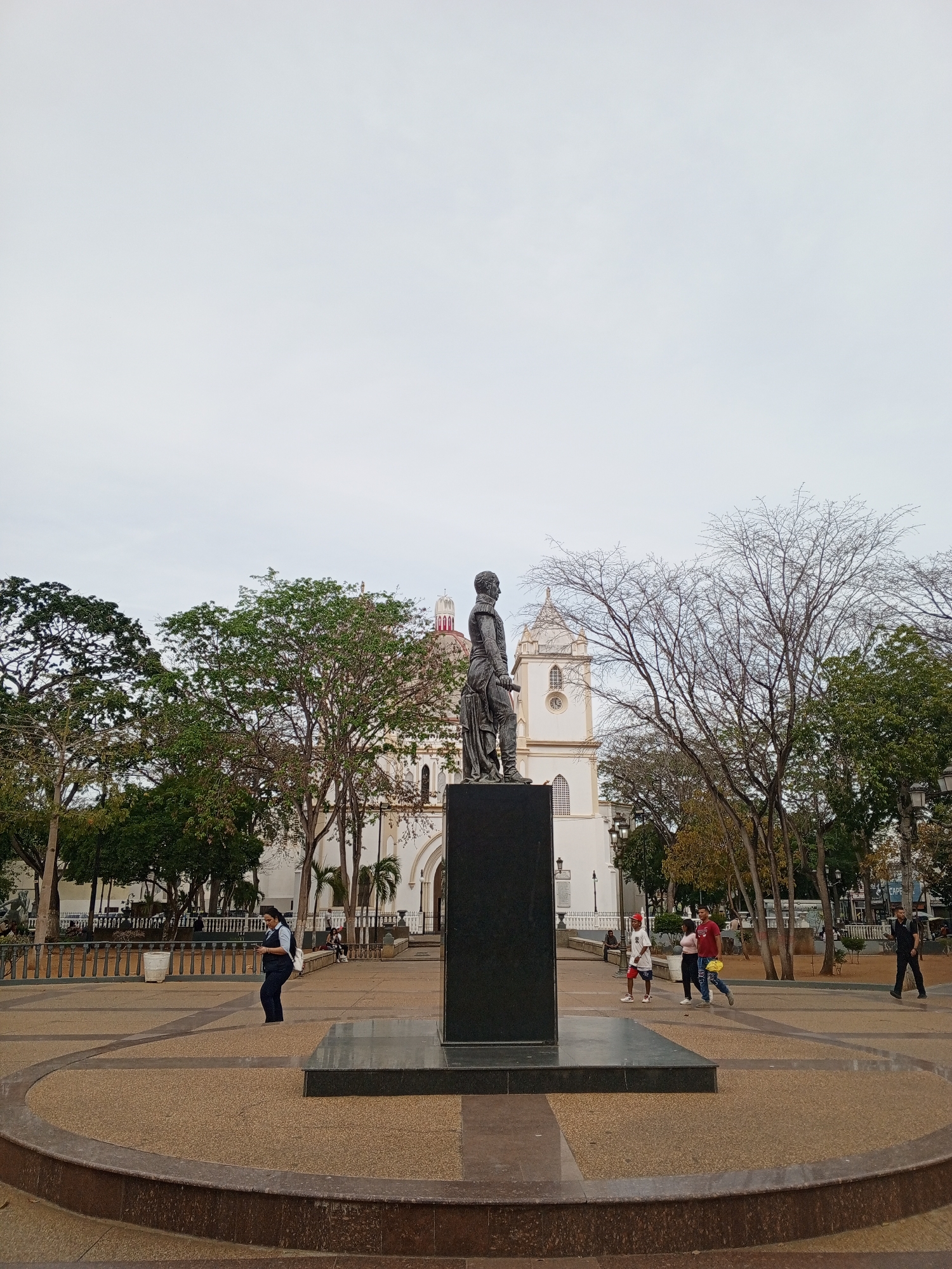 The image shows a statue on a dark rectangular base in a public square, likely in Porlamar, Venezuela. Behind the statue, a white building with a tower and red dome, possibly a cathedral or church, is visible through the trees. Several people are walking around the square, including a woman in a dark suit on the left, a group of three people in the center, and a man in dark clothing on the right. The square has a light brown surface with darker brown accents and a raised circular edge in the foreground. Trees with varying amounts of foliage are scattered around the square. The sky is overcast.