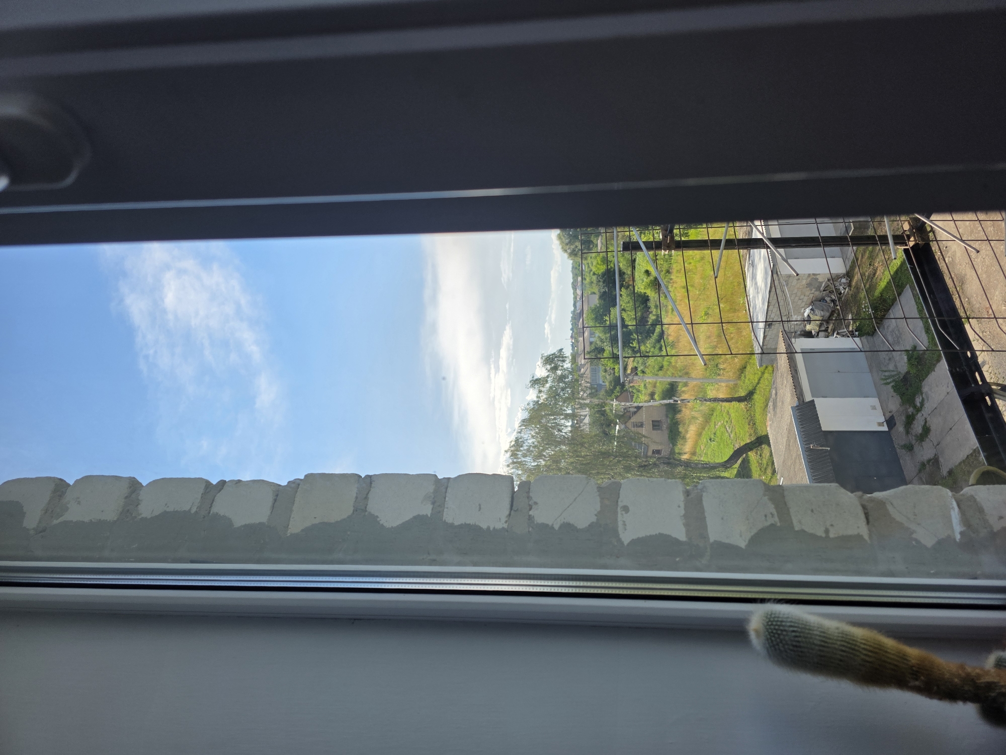 The image shows a view from a window in Dubno, Ukraine. In the foreground, a cactus plant is visible on the windowsill to the left. The window frame is white on the left and dark gray on the right. The window opens outwards, revealing a brick wall with rough cement plaster on the right side of the frame. Beyond the window, a rural or suburban landscape is visible under a bright blue sky with scattered white clouds. The scene includes several small houses, some with tiled roofs, surrounded by green trees and grass. In the middle ground, there are garages with white doors, and a metal fence encloses the property. The overall impression is that of a sunny day in a residential area.