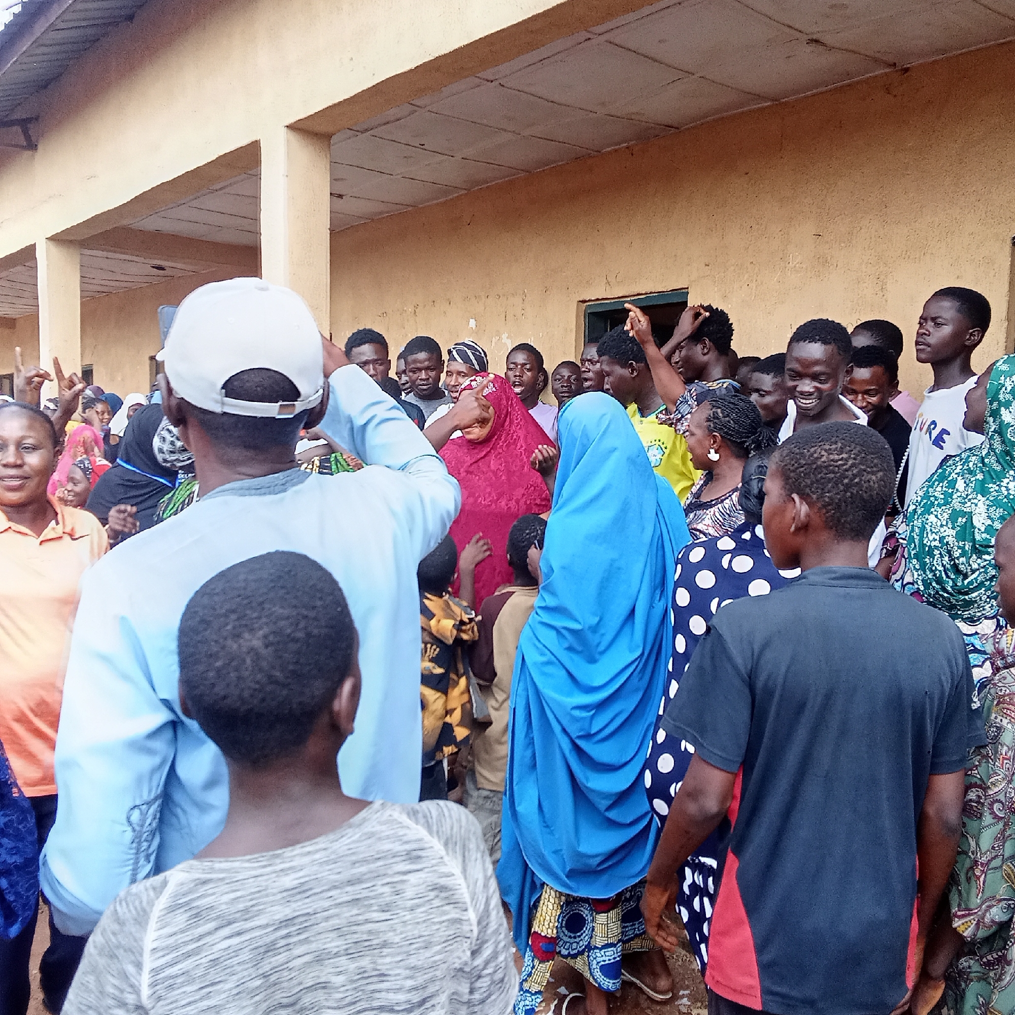A crowd of predominantly black people stands outside a tan building in Akwanga, Nigeria. The crowd includes men, women, and children, many dressed in colorful traditional clothing. Some individuals gesture with their hands raised. The building has a covered walkway supported by pillars. The setting suggests an outdoor gathering or event.