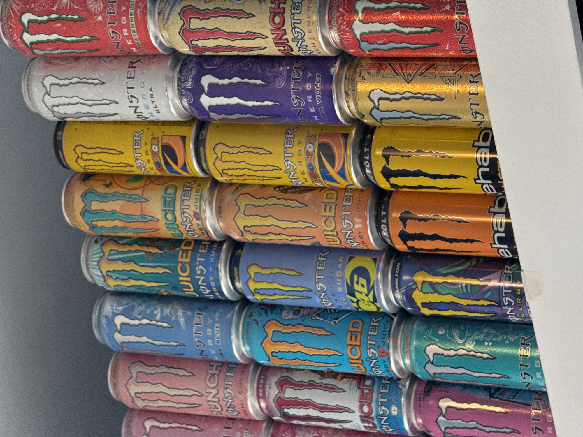 The image shows a stack of Monster Energy drink cans. The cans are stacked on top of each other, displaying a variety of flavors and designs. The flavors include Watermelon, Ultra Violet, Doctor, and others. There is no evidence in the image to suggest this scene is specifically located in Burgas, Bulgaria. There are no people present. The focus is solely on the arrangement and variety of Monster Energy cans.
