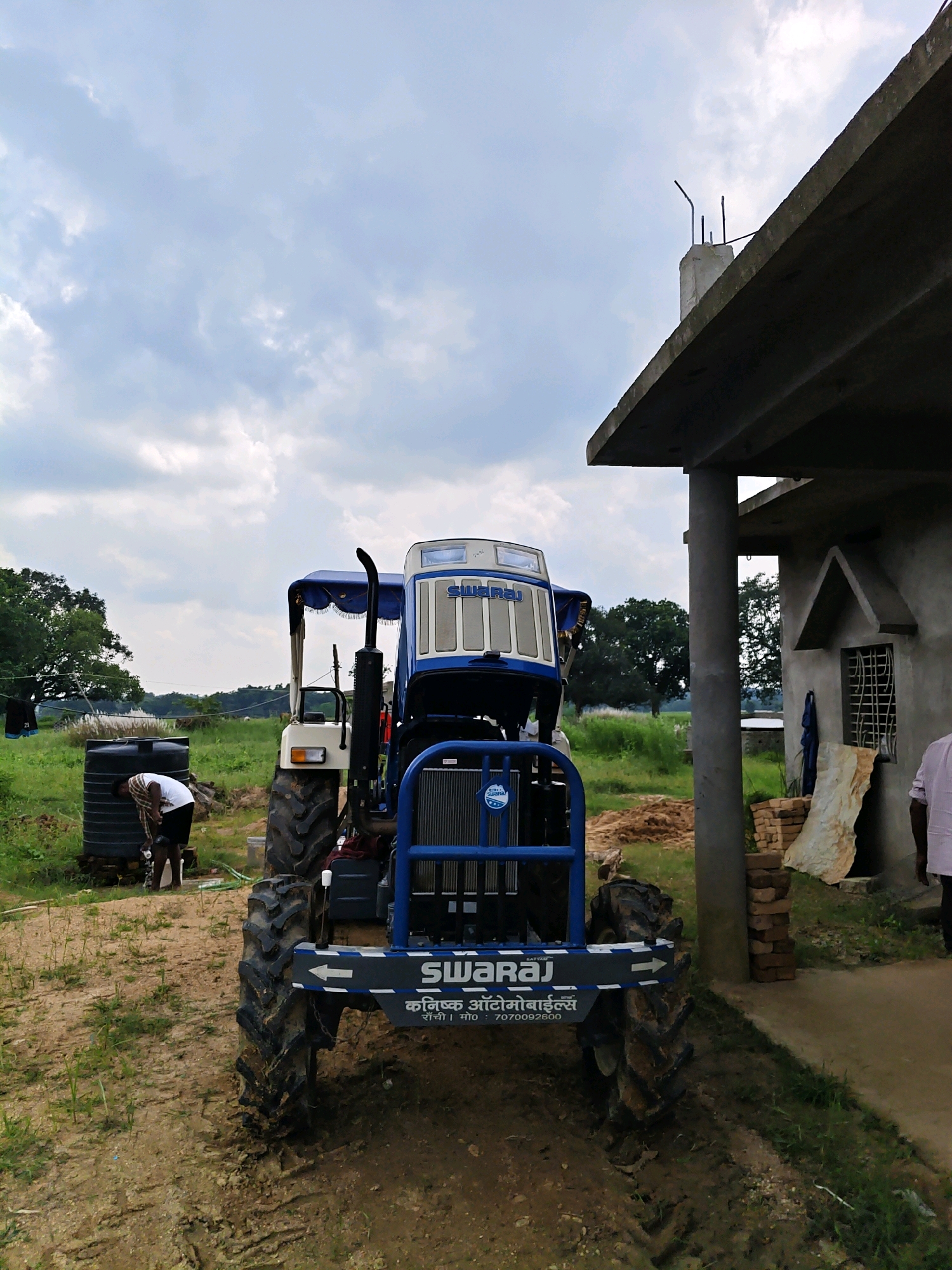 In Sosai, India, a blue and white Swaraj tractor is parked on a dirt road in front of an unfinished concrete building. The tractor's hood is open, revealing its engine. A person is standing next to a large black water tank to the left of the tractor, appearing to be working on something. The scene is outdoors, with green grass and trees in the background under a cloudy sky. The building appears to be under construction, with exposed rebar and stacks of bricks visible.