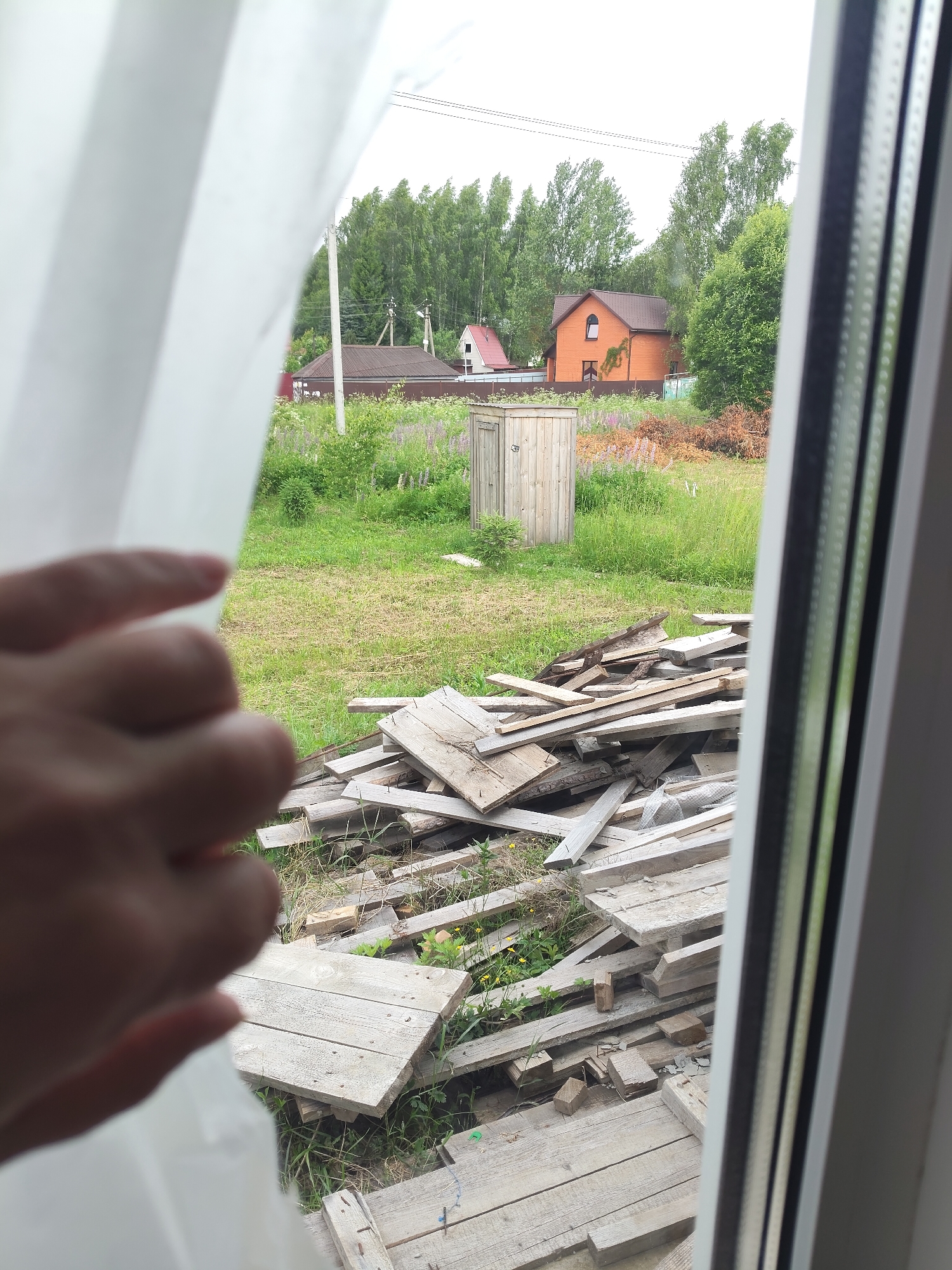 A hand is visible in the foreground, partially obscuring a view through a window. Outside, a yard is filled with a pile of wooden planks, a small wooden outhouse, and various types of vegetation including tall grass and trees. In the background, there are several houses and power lines, suggesting a rural or suburban setting. The sky appears overcast, indicating a cloudy day. The scene appears to be captured from inside a dwelling, looking out. There is no visible text in the image.