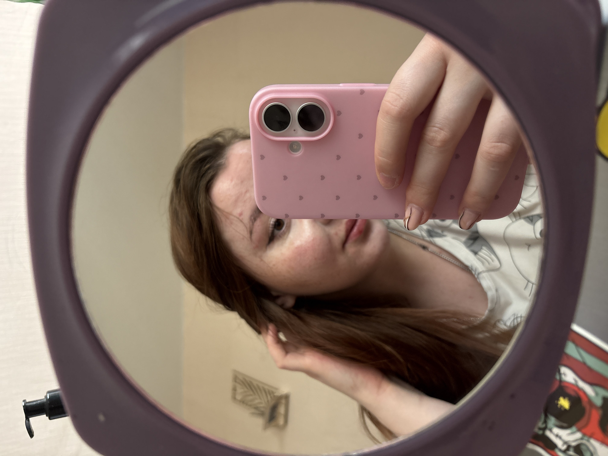 A person is reflected in a round, purple-framed mirror, taking a selfie with a pink smartphone case covered in small hearts. The person has long, brown hair, freckles, and a visible blemish on their forehead. Their fingernails are manicured with a minimalist design. The background reflected in the mirror shows a beige wall with a palm leaf decoration and part of a bed with a white patterned sheet. The scene is indoors. The specific location within Izhevsk, Russia, cannot be determined from the available visual information.
