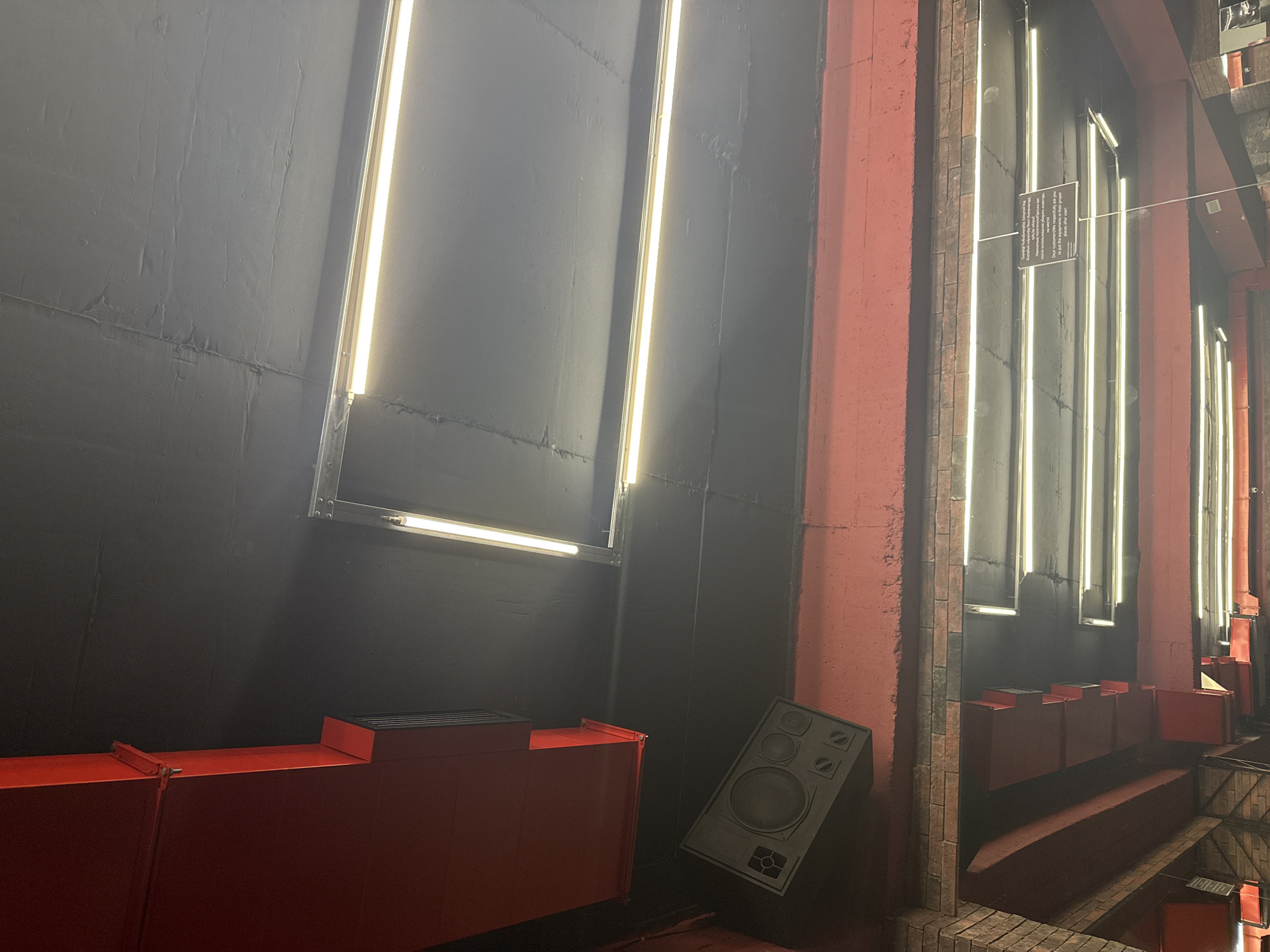 The image shows an interior space with a modern, industrial design. Dominating the scene are vertical, rectangular light fixtures mounted on a dark-colored wall. To the left, a long, red, rectangular structure sits low to the ground, while a speaker leans against the wall near it. The right side of the image reveals a red brick structure with recessed areas and more vertical light fixtures. Signage is attached to one of the fixtures. The floor appears to be constructed from bricks, with glimpses of red elements extending downwards.
Given the text in the signage, it might be a garage in Yerevan, Armenia. The space seems to be arranged for events or performances, considering the lighting and speaker placement.