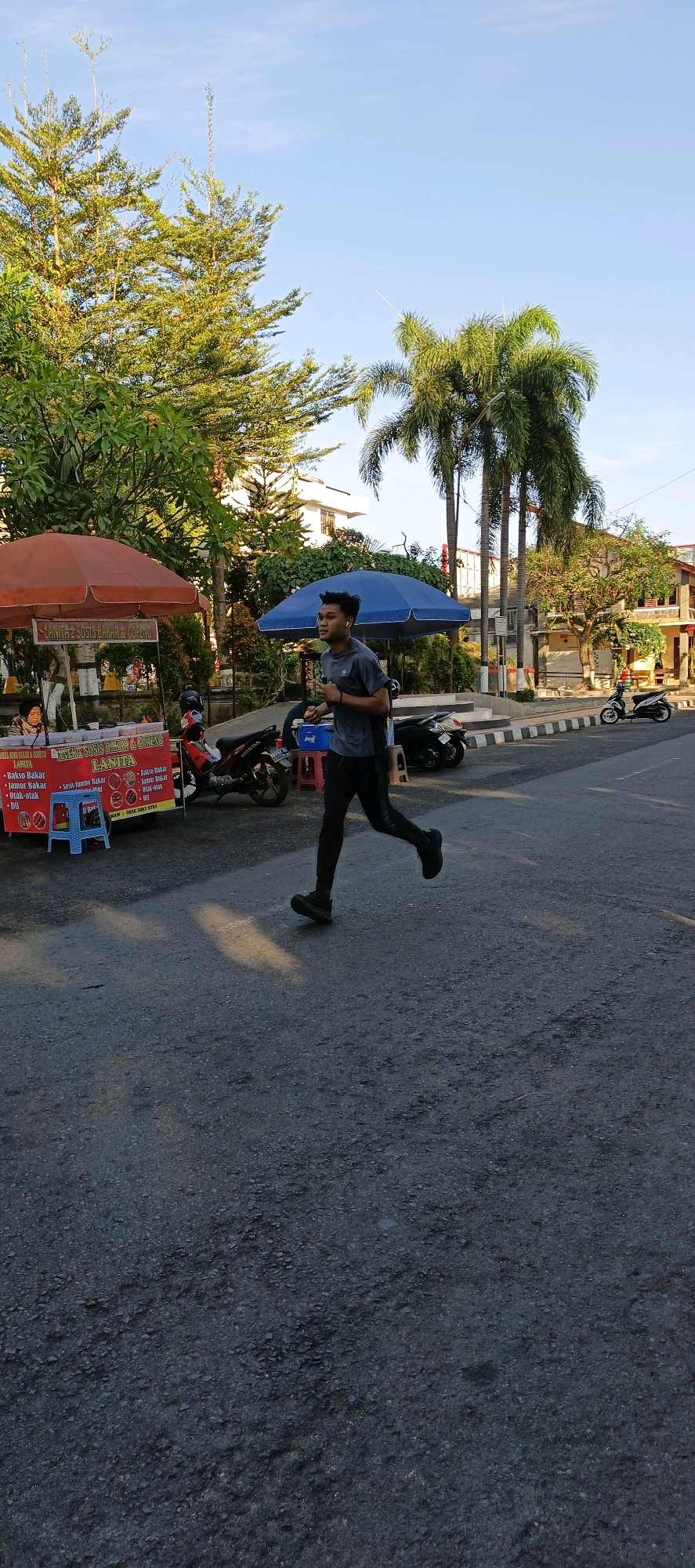 In Klaten, Indonesia, on a paved road with visible asphalt texture, a young man is captured mid-stride while running. He is wearing a dark gray t-shirt and dark pants, with white earbuds in his ears. The scene unfolds outdoors with a clear blue sky above. To the left of the runner, a food stall is set up under an orange awning, displaying menus with Indonesian text and images of food items. Adjacent to the stall, motorcycles are parked. Behind the food stall and further back, a large blue umbrella shades a small area, and more motorcycles are visible. Lush green trees, including palm trees, are scattered throughout the background, with some buildings peeking through the foliage. The overall impression is a snapshot of a casual outdoor scene in an Indonesian city, with the runner being the primary subject of focus in the foreground.