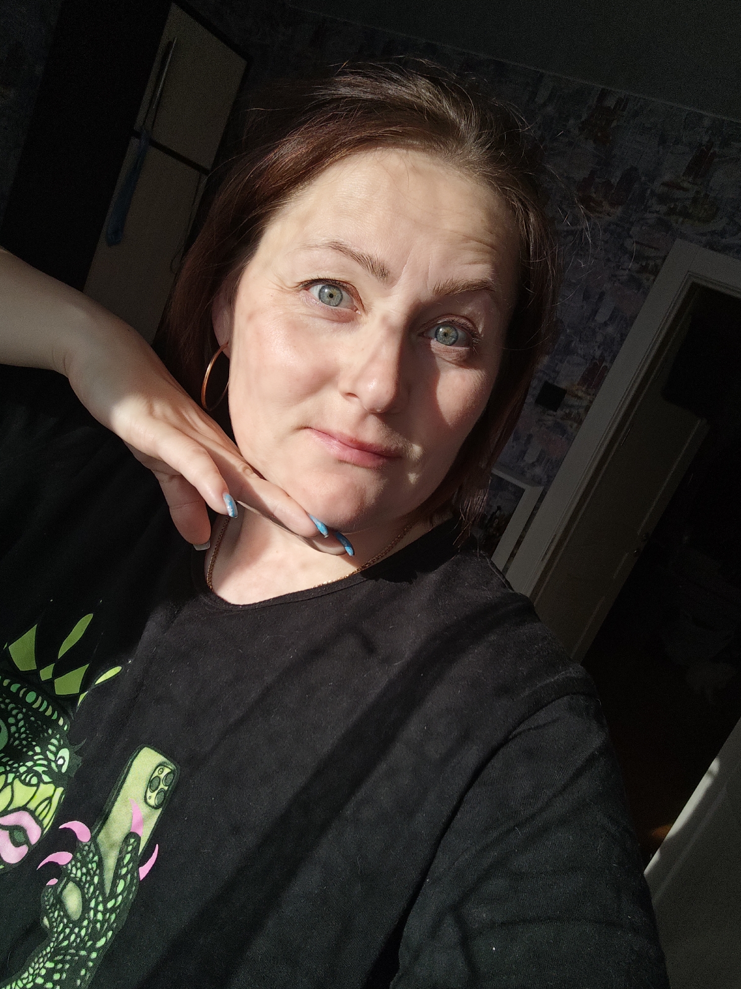 A close-up indoor portrait features a woman with reddish-brown hair and green-blue eyes, looking directly at the camera with a subtle smile. Strong, directional sunlight illuminates the right side of her face and her raised left hand, creating clear shadows. She wears a black t-shirt with a vibrant, stylized graphic design on the left side, depicting a green, monster-like character with pink accents, holding a light green smartphone with a triple camera. A thin gold chain necklace and a gold hoop earring are visible. Her left hand, adorned with light blue manicured nails, gently rests near her jawline and neck. The background is dimly lit, revealing dark patterned wallpaper on the right and a very dark area on the left with a hint of a light-colored object, possibly a cabinet or mirror, and a blue cloth. The natural sunlight suggests it is daytime.