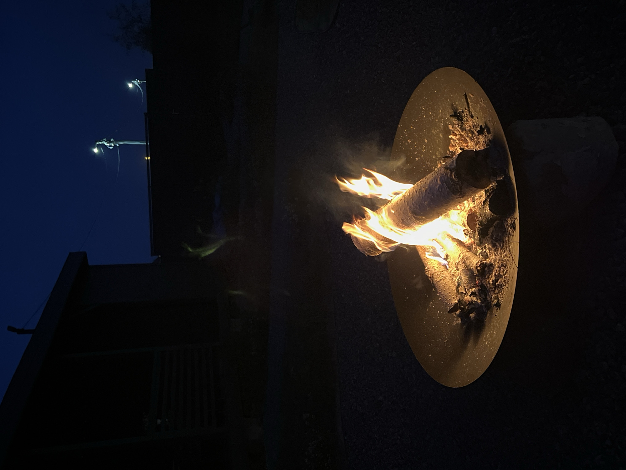 The image depicts a fire pit with burning logs in the foreground, set against a dark, twilight sky. A portion of a building with horizontal siding is visible on the left, silhouetted against the sky. In the background, two streetlights on poles illuminate the scene. The ground around the fire pit appears to be covered in gravel. The overall atmosphere is serene and dark. The location is identified as Великие Озера Кп, Russia.