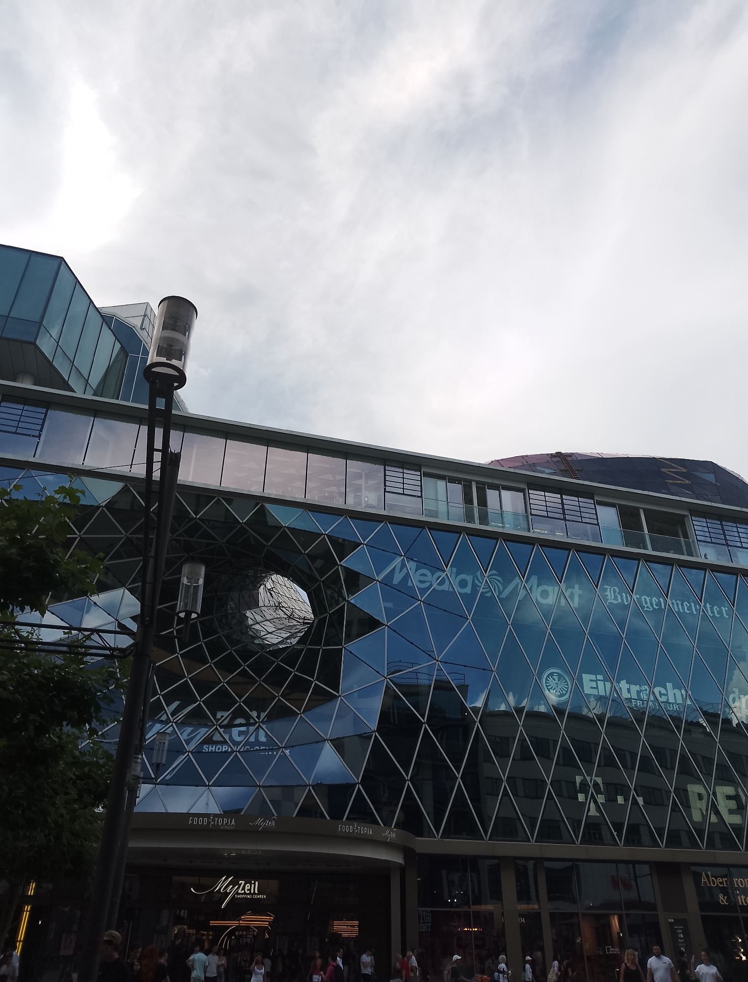 The image shows a street-level view of the MyZeil shopping center in Frankfurt, Germany. The modern glass-facade building features a distinctive geometric pattern. Reflections on the glass reveal other buildings and the sky. A tall lamppost with multiple light fixtures is visible in the foreground. Pedestrians are seen on the sidewalk in front of the shopping center entrance, which is illuminated. Signs for "MyZeil SHOPPING CENTER," "FOOD TOPIA," and "Eintracht FRANKFURT" are visible. The sky above is overcast with white clouds.