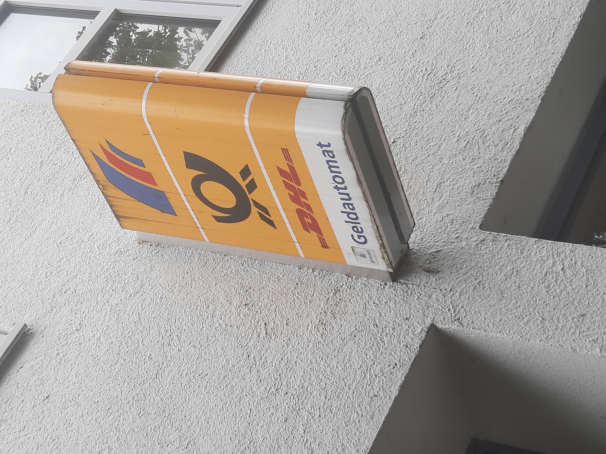 The image shows a weathered, yellow and orange sign for "Post und DHL Geldautomat" mounted on the textured white exterior wall of a building. The sign, which is rectangular and appears to be illuminated from within, is angled outwards from the wall. At the top of the sign, a logo featuring red, blue, and white stripes is visible, followed by a large, dark brown Post horn logo. Below this, the red DHL logo is prominent. At the bottom of the sign, in blue text, is "Geldautomat" and a small "girocard" logo. To the right of the sign, a window with white framing is visible, reflecting trees and sky. The building's wall has a rough, uneven texture.