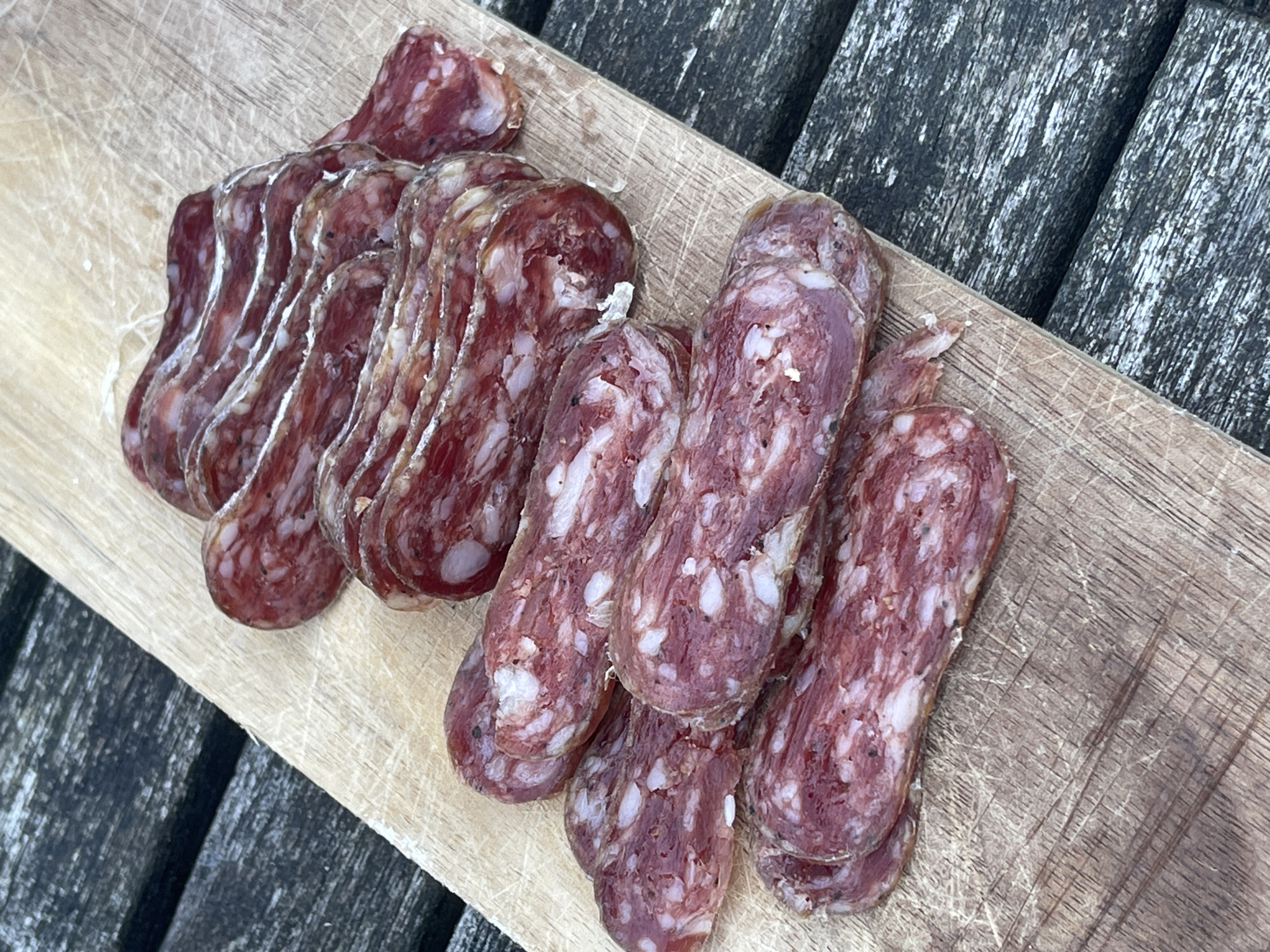 The image shows a wooden cutting board placed on a dark, weathered wooden surface. Arranged on the board are two piles of sliced sausage or salami. The slices in the first pile are thinly cut and have a lighter-colored fatty edge. The slices in the second pile are thicker and show more white marbling throughout the meat. The location is unknown.