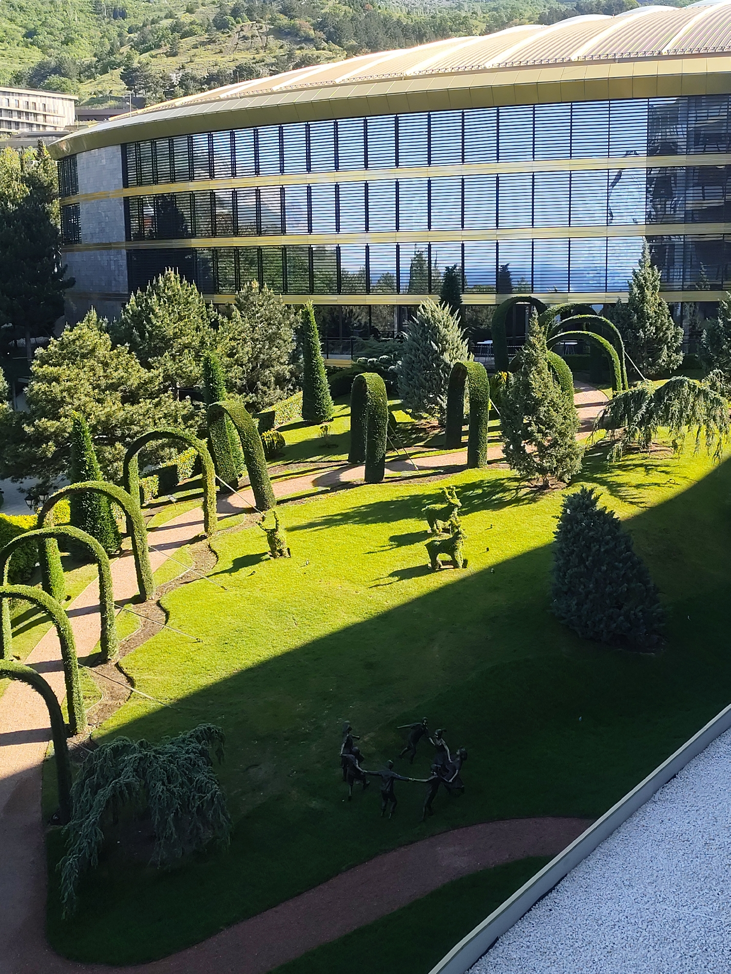 The image depicts a manicured garden adjacent to a modern building, likely located in Unknown, Unknown. The building has a multi-story facade with horizontal windows and a curved roofline.
The garden features a variety of topiary, including archways, cylinders, and animal shapes (deer). There are also standard trees and shrubs arranged throughout the garden. In one area, there is a bronze sculpture of multiple figures holding hands.
A narrow path winds through the garden. A distinct shadow line cuts across the lawn, indicating a strong light source, presumably the sun. The garden appears well-maintained.