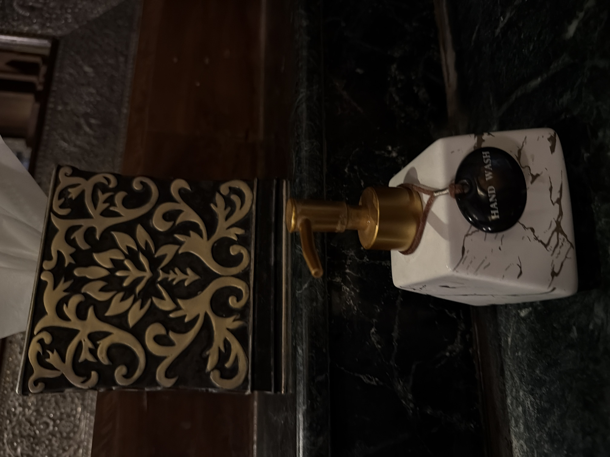 A close-up shot shows a decorative tissue box with a black and gold ornate pattern, filled with white tissues, positioned behind a white and gold marble-patterned hand wash dispenser. The dispenser has a gold pump and a small, dark circular tag that reads "HAND WASH." Both items are placed on a dark, glossy surface that appears to be marble. The background is dimly lit with dark wooden elements and a textured wall. The scene is indoor, likely in a bathroom or washroom. The image appears to have been taken in Ko Samui District, Thailand.