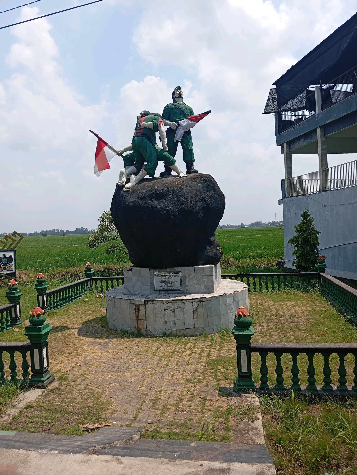 The image depicts a memorial statue in Leman Benawi, Indonesia. The statue, situated in front of a building with a dark roof and a balcony, features three figures dressed in green military uniforms. Two figures are actively raising an Indonesian flag (red and white), while a third stands nearby. The figures are positioned atop a large, dark boulder that rests on a white, multi-tiered base. A plaque is visible on the base of the monument. The surrounding area is an open space with a paved walkway and green fencing with decorative elements. Beyond the memorial, lush green rice fields stretch into the distance under a partly cloudy blue sky. The scene appears to be a historical monument commemorating a significant event in Indonesian history, likely related to independence or military struggle.