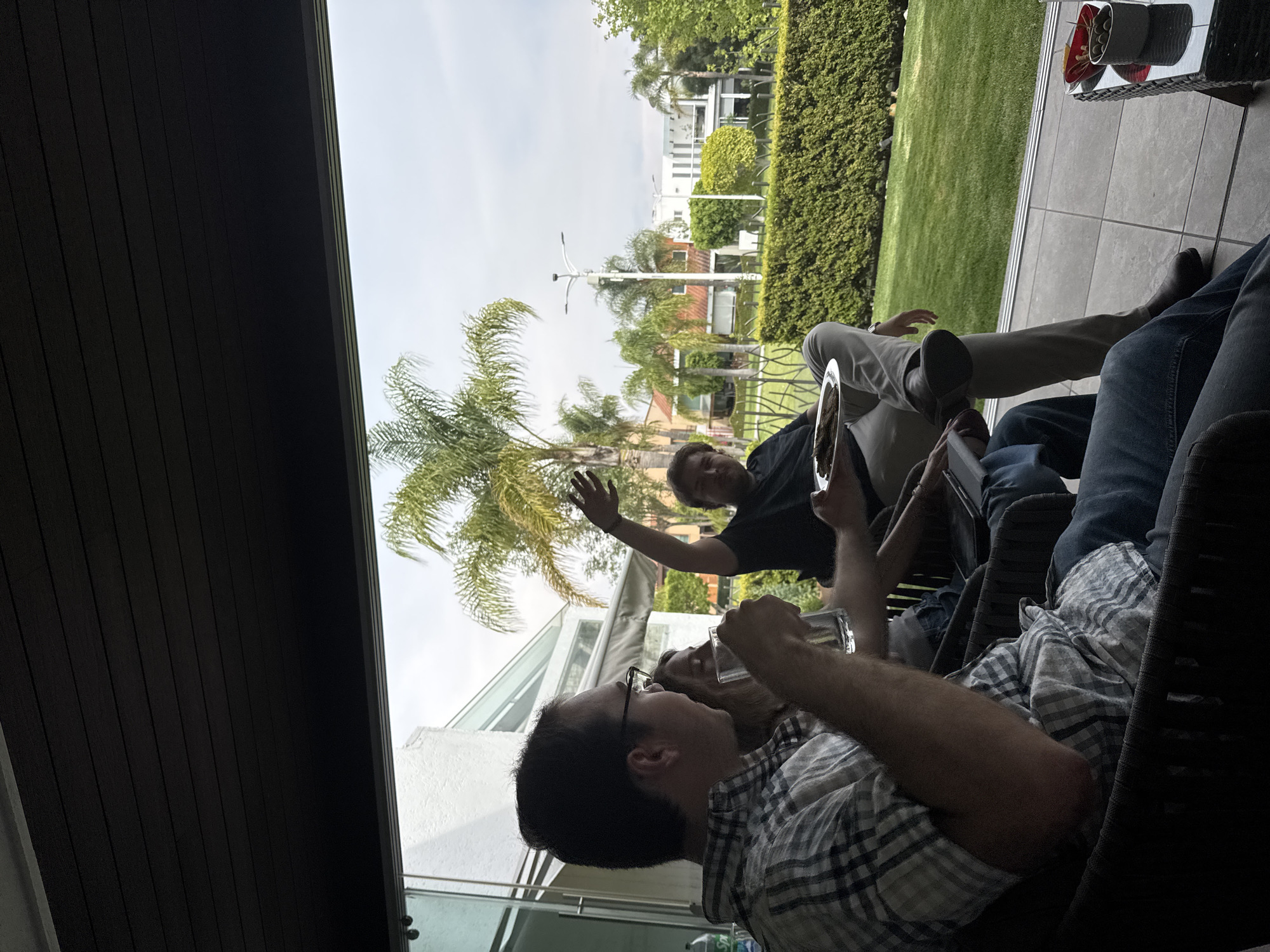 The image shows a group of people gathered on a patio. One man is wearing glasses and a plaid shirt and holding a glass. Another man is raising his hand. Other people and a building are partially visible in the background. The setting appears to be outdoors, with greenery visible in the distance. The location is unknown, and there is no information available to indicate that the image was taken in San Bernardino Tlaxcalancingo, Mexico.