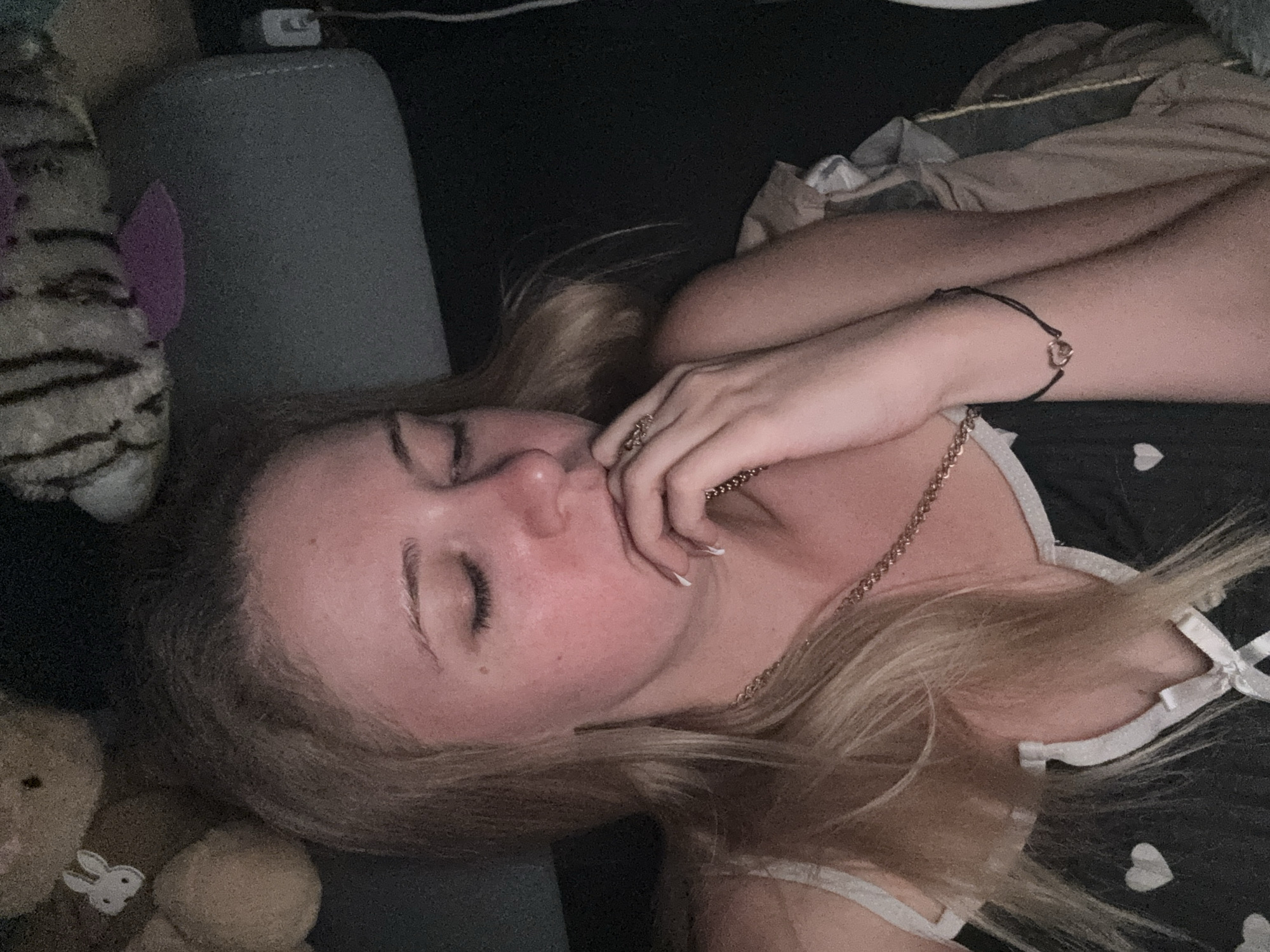 A young woman with blonde hair is lying down, eyes closed, with her hand near her mouth. She is wearing a black camisole with white hearts and a white trim, and a gold chain necklace. A black bracelet with a small heart charm is visible on her wrist. She appears to be in a dark room, possibly a bedroom, with stuffed animals visible in the background. A gray headrest is behind her, and some cables are plugged into a power outlet on the right side of the image.