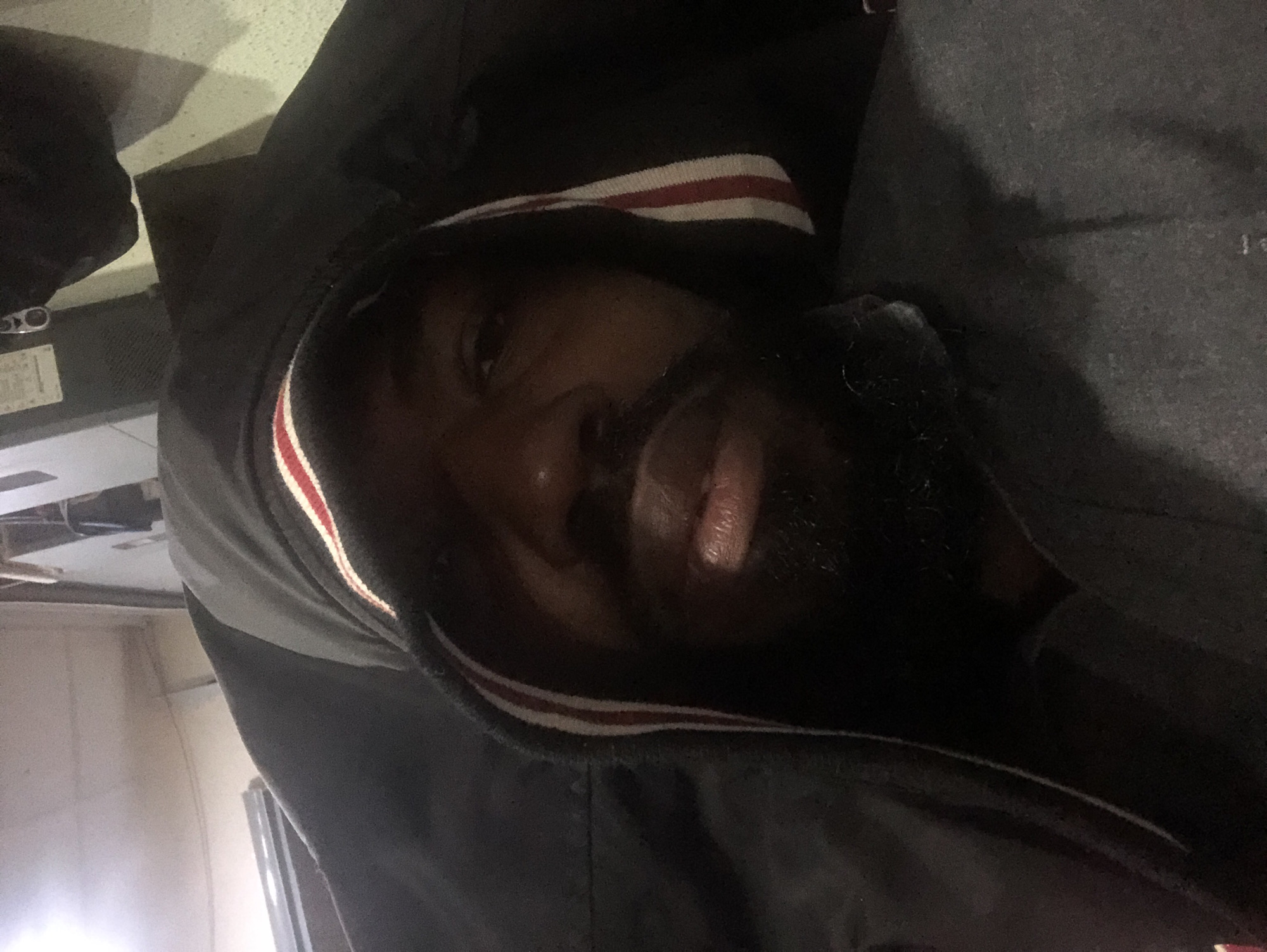A medium shot photograph shows a Black man from the chest up, looking directly into the camera. He is wearing a dark hooded jacket, with the hood pulled up over his head. The jacket has a red and white striped trim along the edge of the hood. The man has a beard and his face is partially obscured by shadows and the hood. The background appears to be an indoor setting with what looks like industrial equipment and some lighting fixtures on the ceiling. The scene is dimly lit. There is no indication of the specific location within Ikeja, Nigeria.