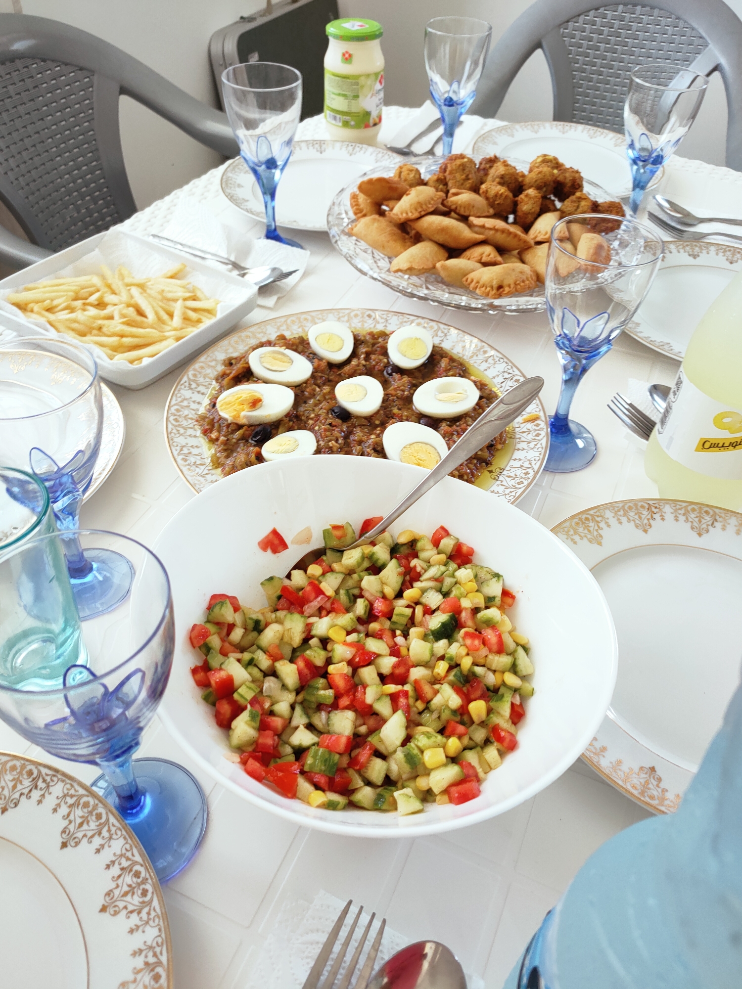 The image depicts a table set for a meal, likely in a domestic setting in Mrezga, Tunisia. A spread of various dishes is laid out on a white tablecloth. In the foreground, a white bowl contains a chopped salad composed of diced tomatoes, cucumbers, and corn kernels, with a serving spoon resting inside. To the left, a clear glass with a blue stem and base holds a portion of the salad. Partially visible to the left is another similar glass.
Further back on the table, a white rectangular dish holds a generous portion of golden french fries. Adjacent to this is a clear glass plate filled with an assortment of fried pastries and small, dark-colored fried balls. Another plate, decorated with a gold and white pattern, is visible in the background to the right, as is a partially obscured white serving dish with a gold rim.
A rectangular dish containing a spread with sliced hard-boiled eggs and what appears to be olives is situated prominently. Several blue-stemmed glasses are arranged around the table. In the background, a jar with a green lid and label is present. A bottle of a yellow beverage, possibly lemonade, with Arabic text on its label, stands on the right side of the table. A fork and knife are visible on a napkin near the beverage. The chairs at the table are dark gray and appear to be made of plastic. The overall impression is of a casual meal gathering.