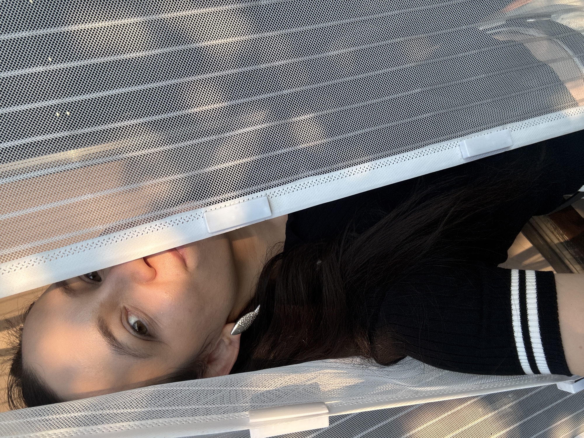 A woman is partially visible behind a white mesh screen, with sunlight casting shadows across her face and shoulder. She has long dark hair and is wearing a black top with white stripes on the sleeves. She is also wearing a fish-shaped earring on her left ear. The scene appears to be indoors, possibly near a window or door, with the mesh screen serving as a barrier. The environment suggests a domestic setting. There is no indication of the city of Chaikovsky, Russia, in the image itself.