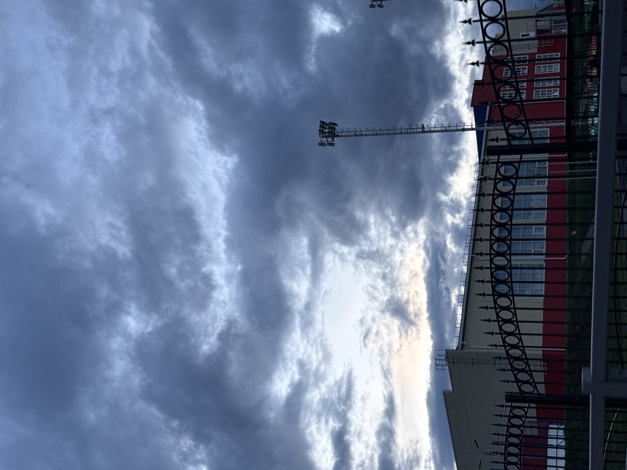 The image shows a cloudy sky above a building and a metal fence. The building is red and white, with multiple windows. A tall metal light tower is visible in the background, partially obscured by clouds. The metal fence in the foreground is black with decorative circles and spikes. The location is Vereshchagino, Russia.