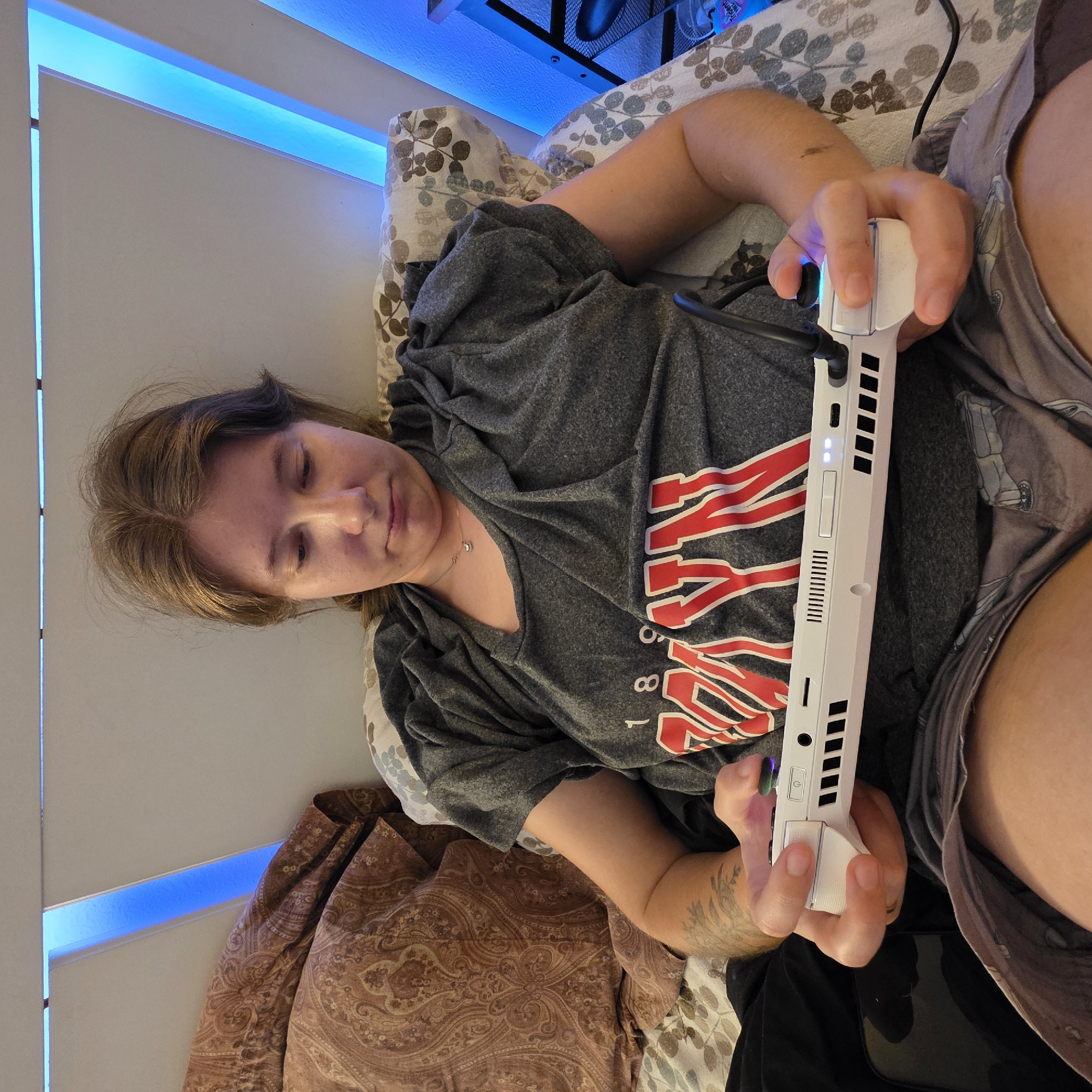 A person is playing a white handheld gaming console while lying in bed. The person is wearing a gray t-shirt with a red and white graphic design on it and gray shorts. They have brown hair and are looking down at the console. The bed has a patterned comforter and several pillows. Blue LED lights are visible behind the person, illuminating the wall. The scene appears to be in a bedroom. No specific location in Portland, United States of America, can be determined from the image.